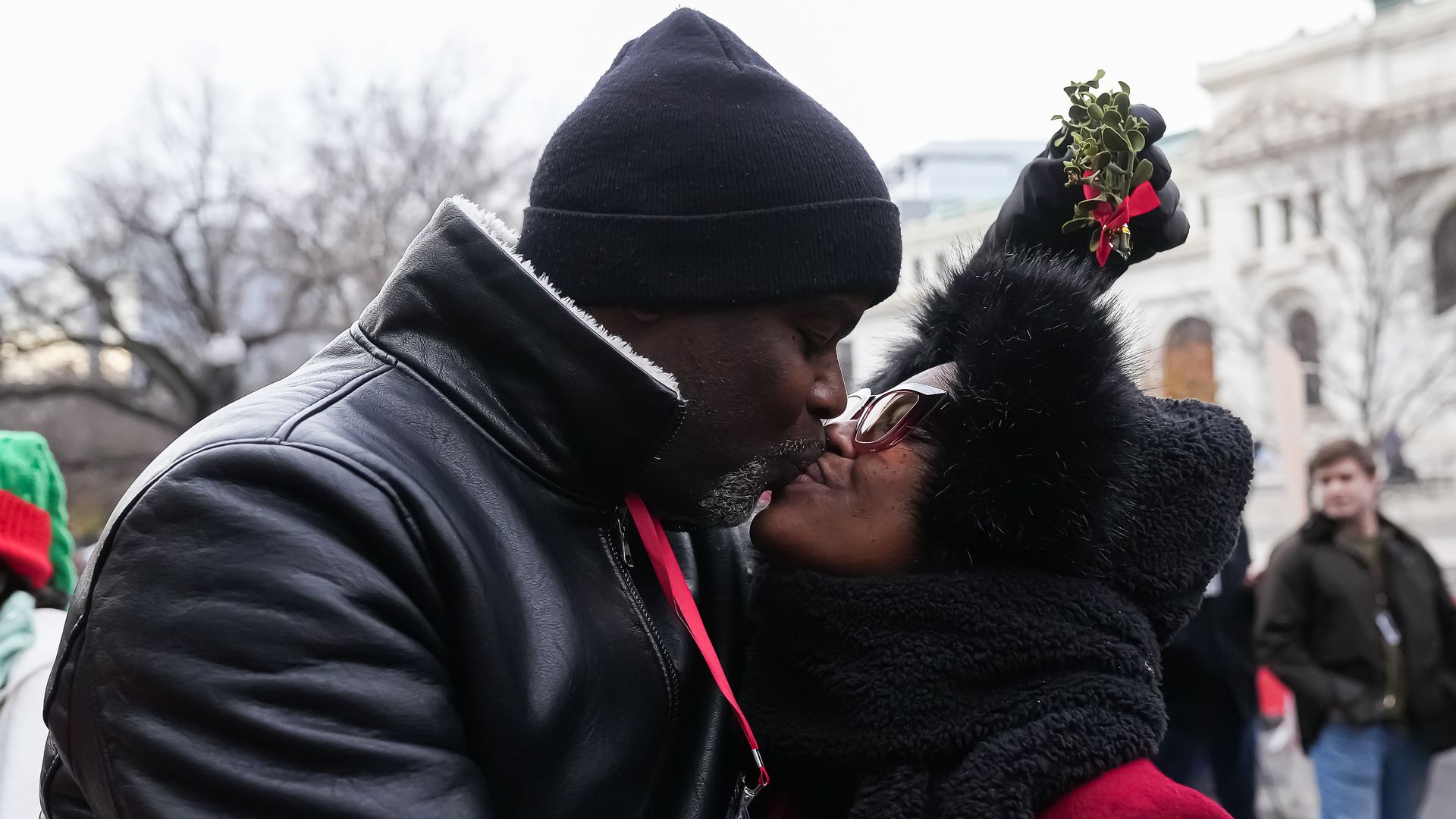 A couple dressed in winter clothing share a kiss outdoors, the woman holding mistletoe above them with a red bow. They wear black hats and coats; bare trees and buildings are in the background.