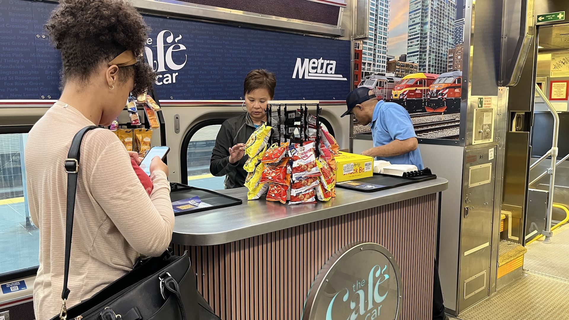 A woman with a phone and black bag stands at a snack counter inside a train labeled "the Café car" with racks of snacks; two workers attend to the counter in front of a cityscape poster.