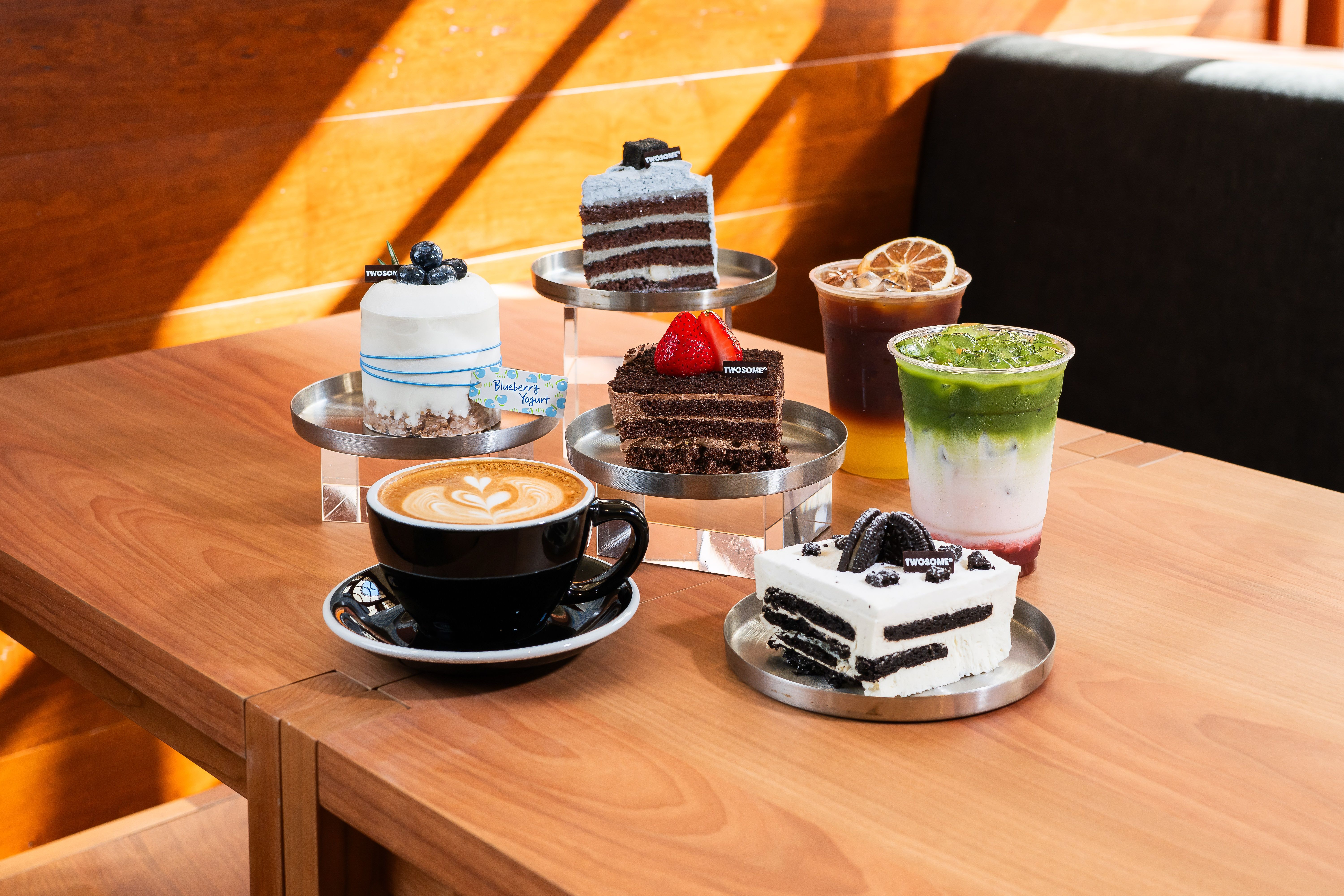 Tiered metal stands hold a white frosted cake with blueberries and a chocolate-layer cake with strawberry; foreground shows black coffee with latte art and a green iced drink on a sunlit table.