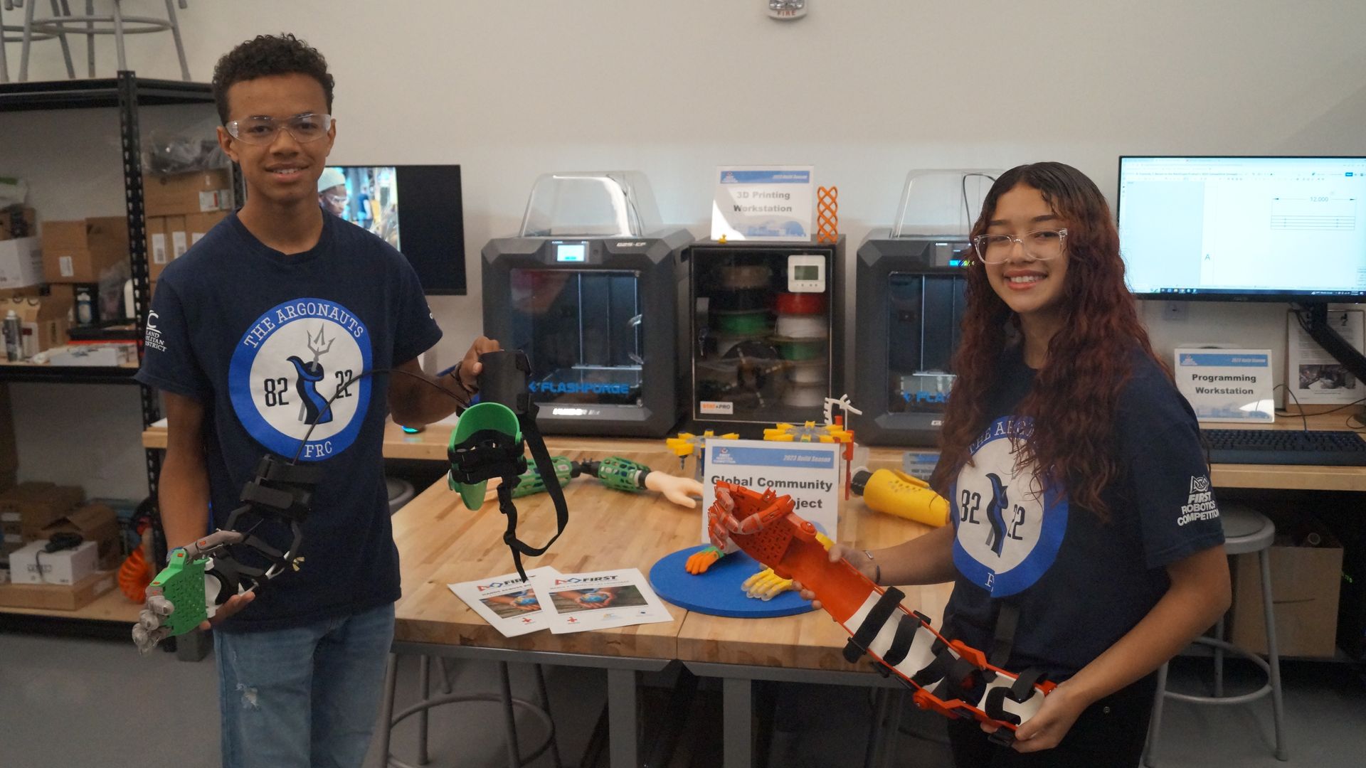 Students in navy t-shirts hold up 3D-printed prosthetic limbs. 