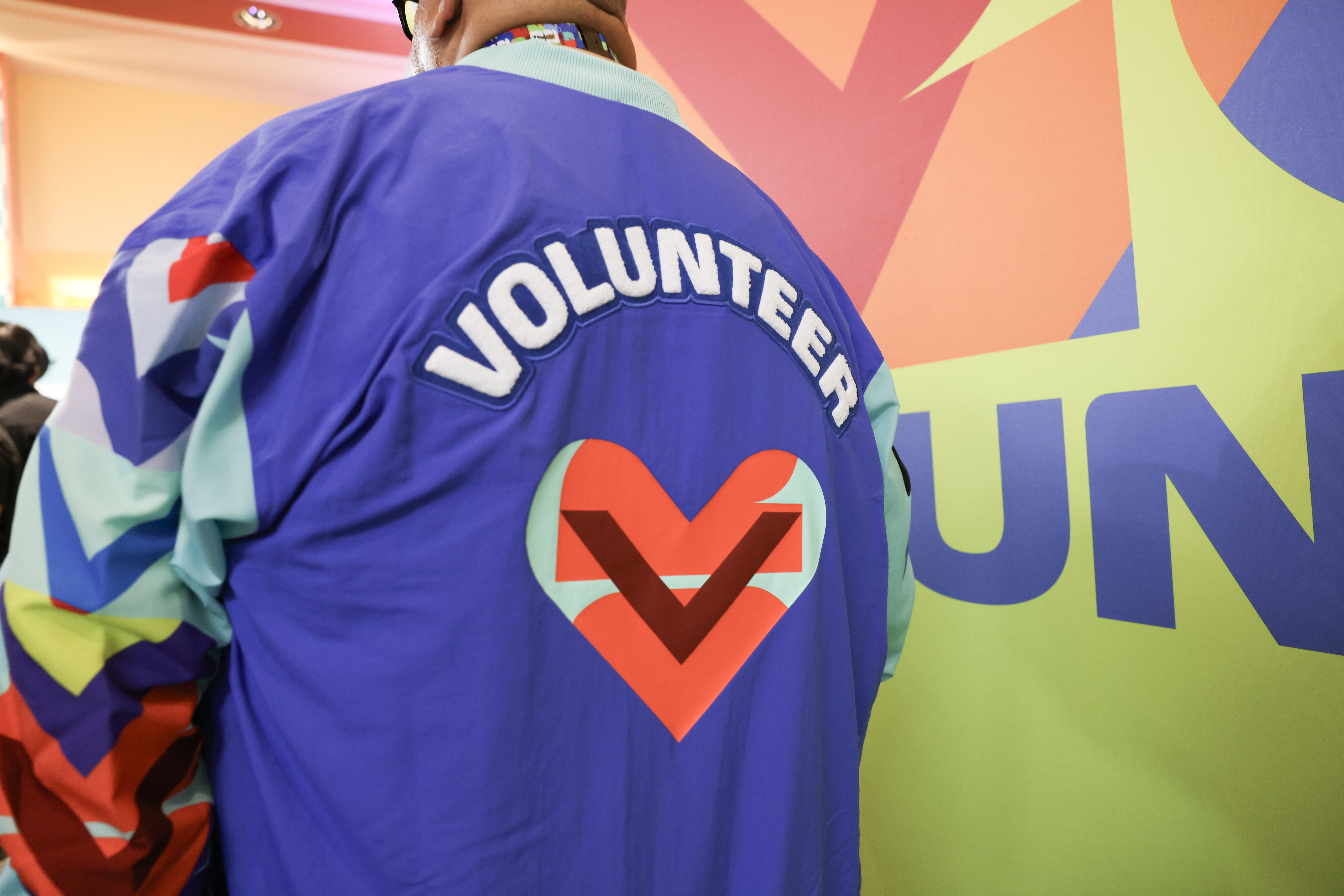 Back of a person wearing a blue jacket with white VOLUNTEER on the back, colorful sleeves; a vivid geometric mural in pink, orange, yellow and green in the background.