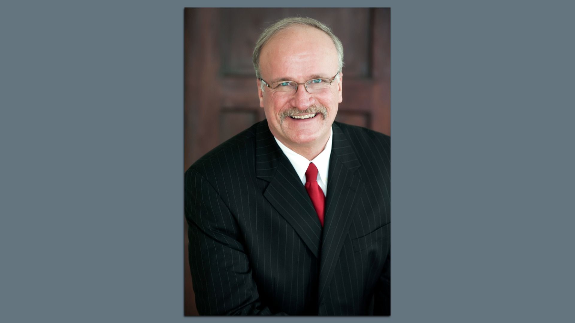 Portrait of a smiling middle-aged man with glasses and a mustache wearing a dark pinstripe suit, white shirt, and red tie, posed against a wooden background.