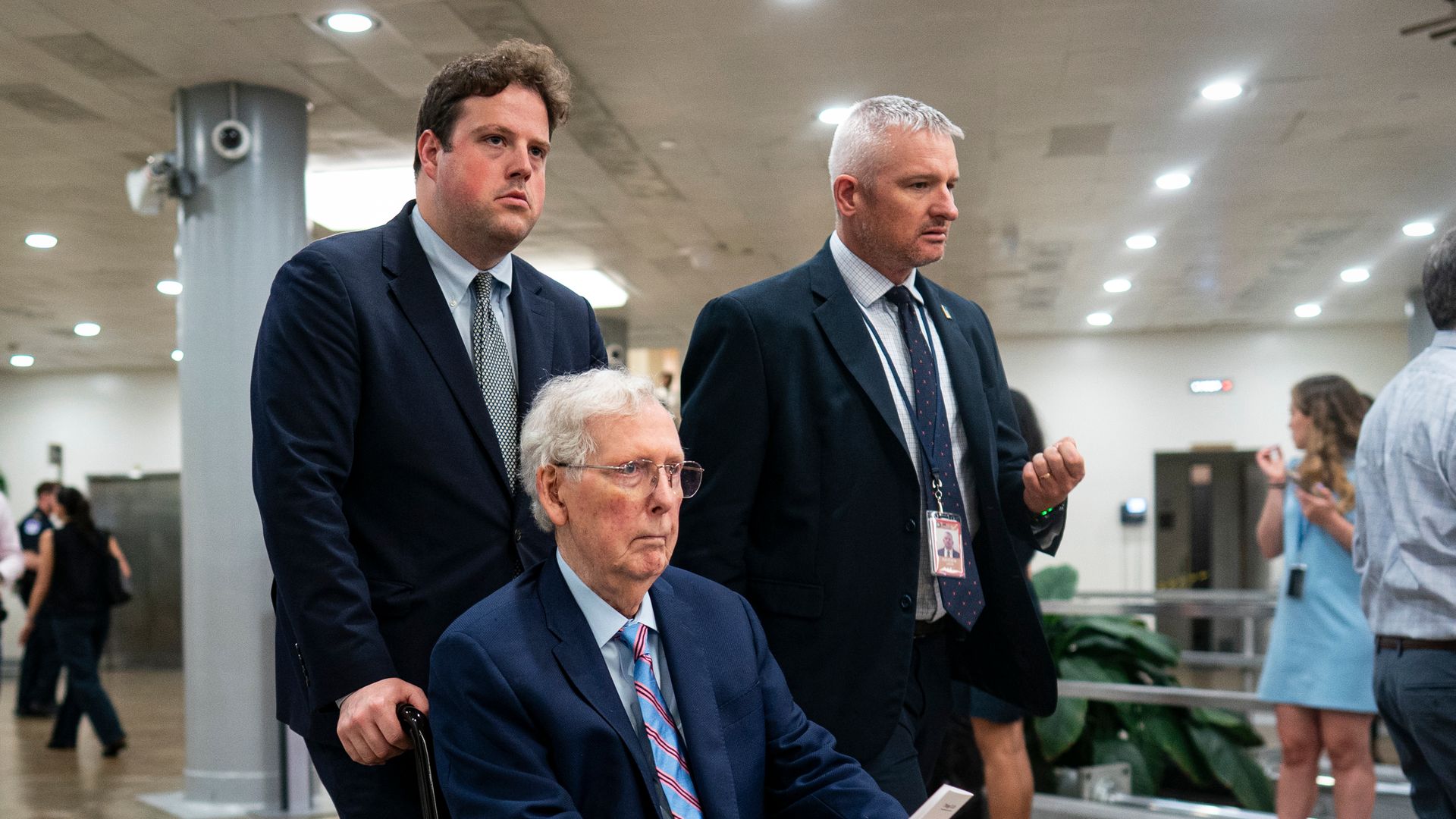 Two men in suits walk through a hallway while one of them pushes an older man, Sen. Mitch McConnell, in a wheelchair.
