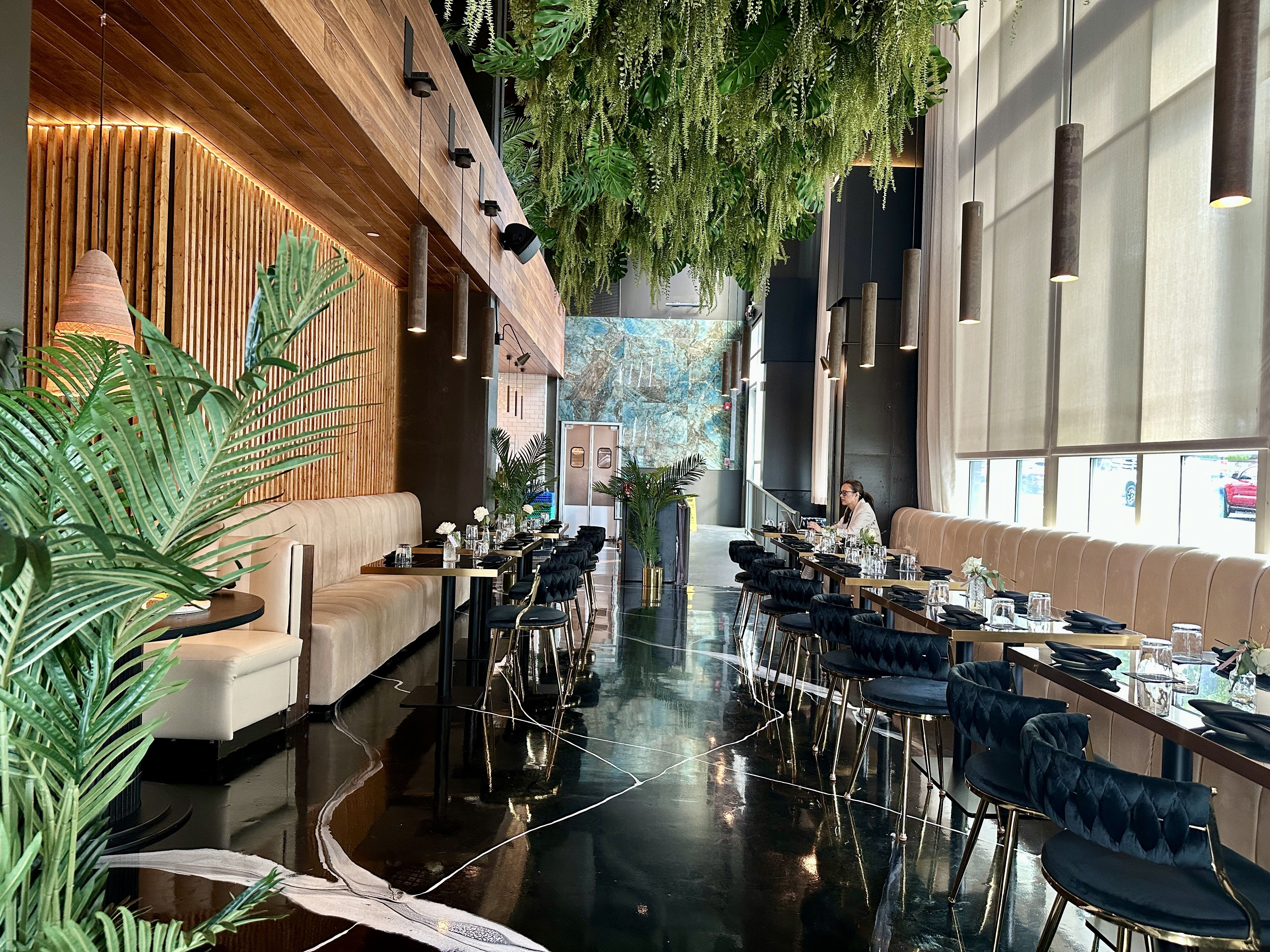 LILA. Indoor restaurant with beige booths along a wooden slat wall, hanging greenery overhead, black glossy floor, and pendant lights. Tables are set with glassware; a person sits at the far end.