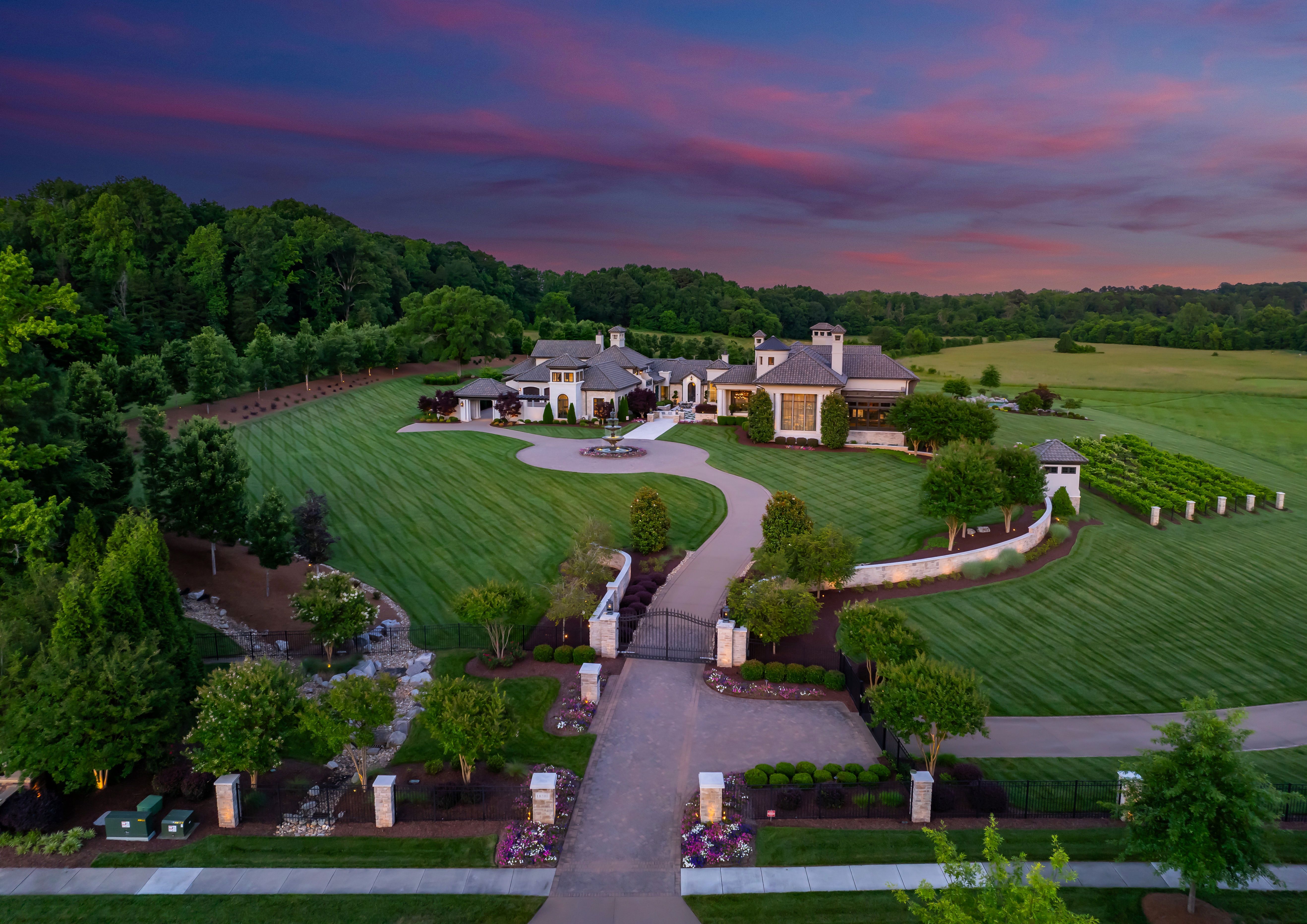 A large, elegant estate with a gated entrance, circular driveway, green manicured lawn, surrounded by trees and fields under a colorful purple and pink sunset sky.