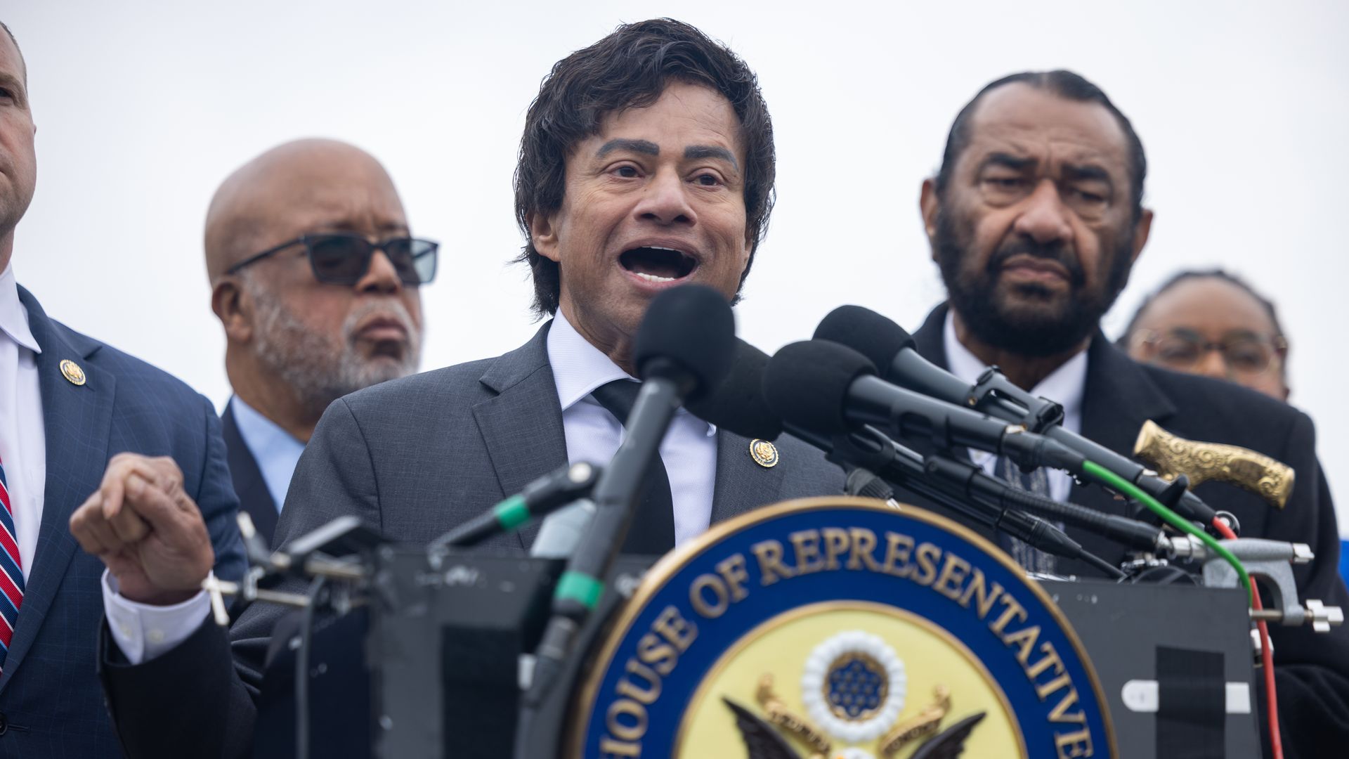 Man in a gray suit speaks at a podium with multiple microphones; a U.S. House of Representatives seal is visible, flanked by men in suits at a press conference.