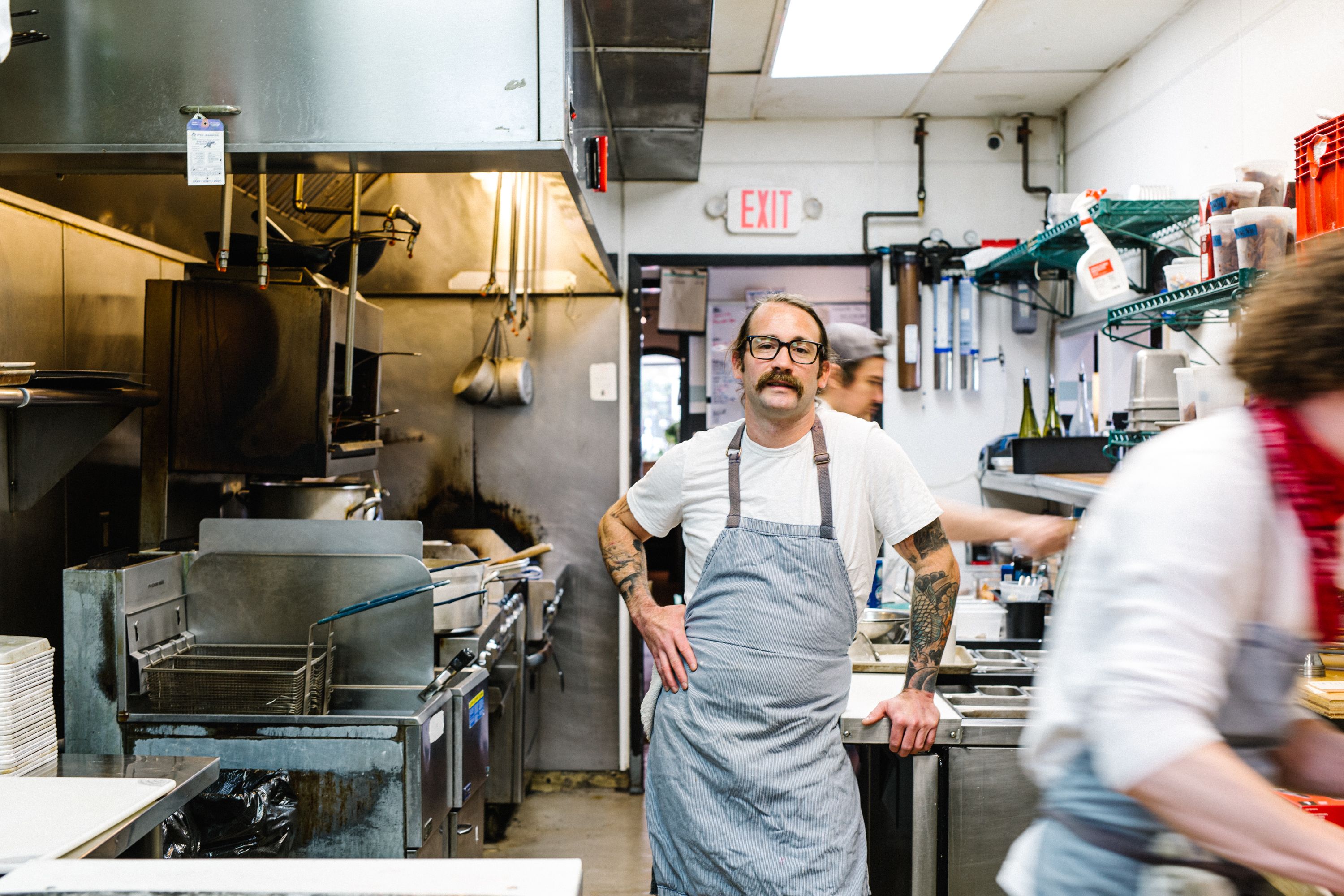 A man with a mustache stands in a busy kitchen. 