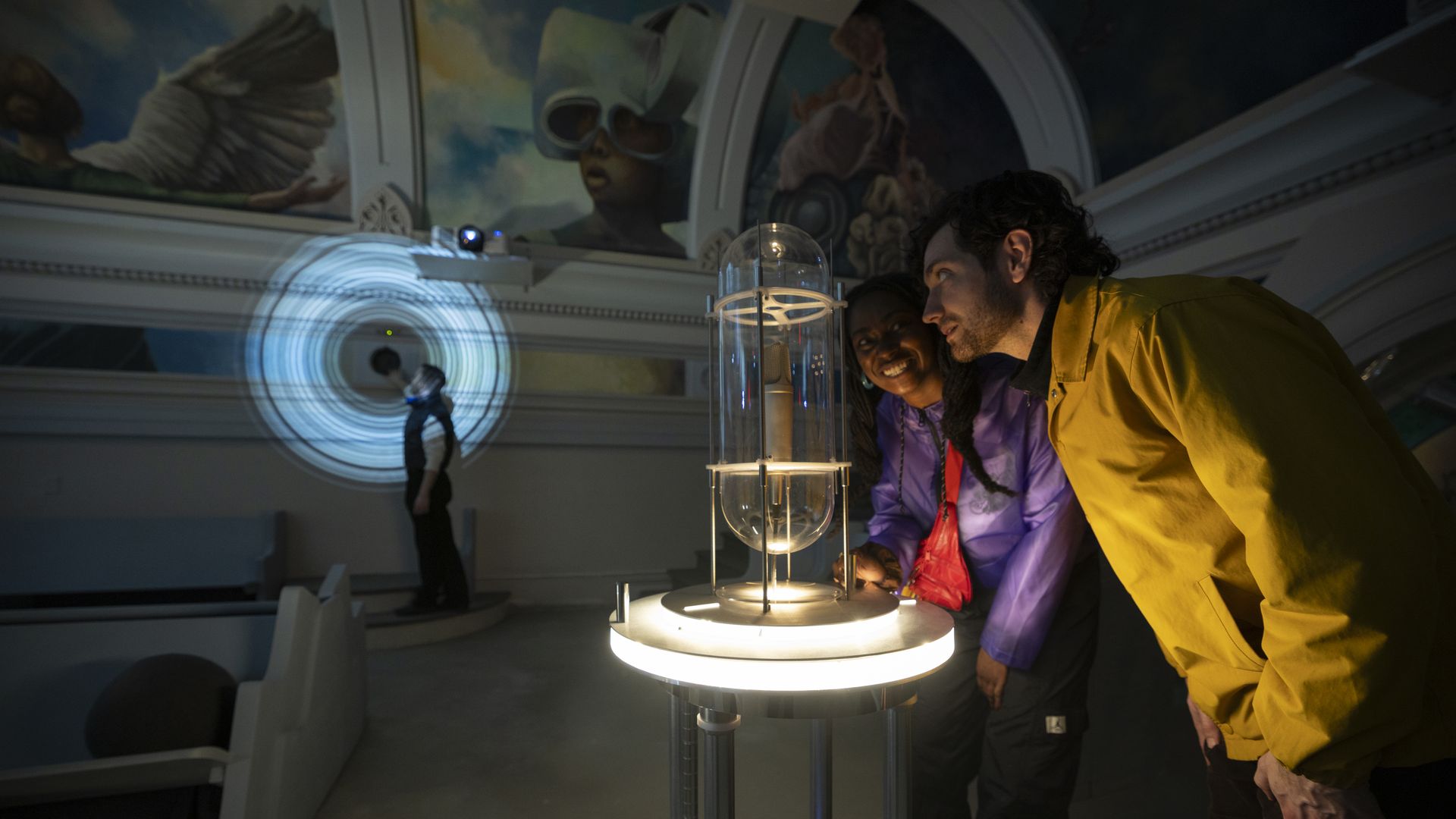 Two visitors, a man in a yellow jacket and a woman in purple, lean over a glass capsule on a glowing pedestal as a blue ring projection and mural loom in the dim museum.