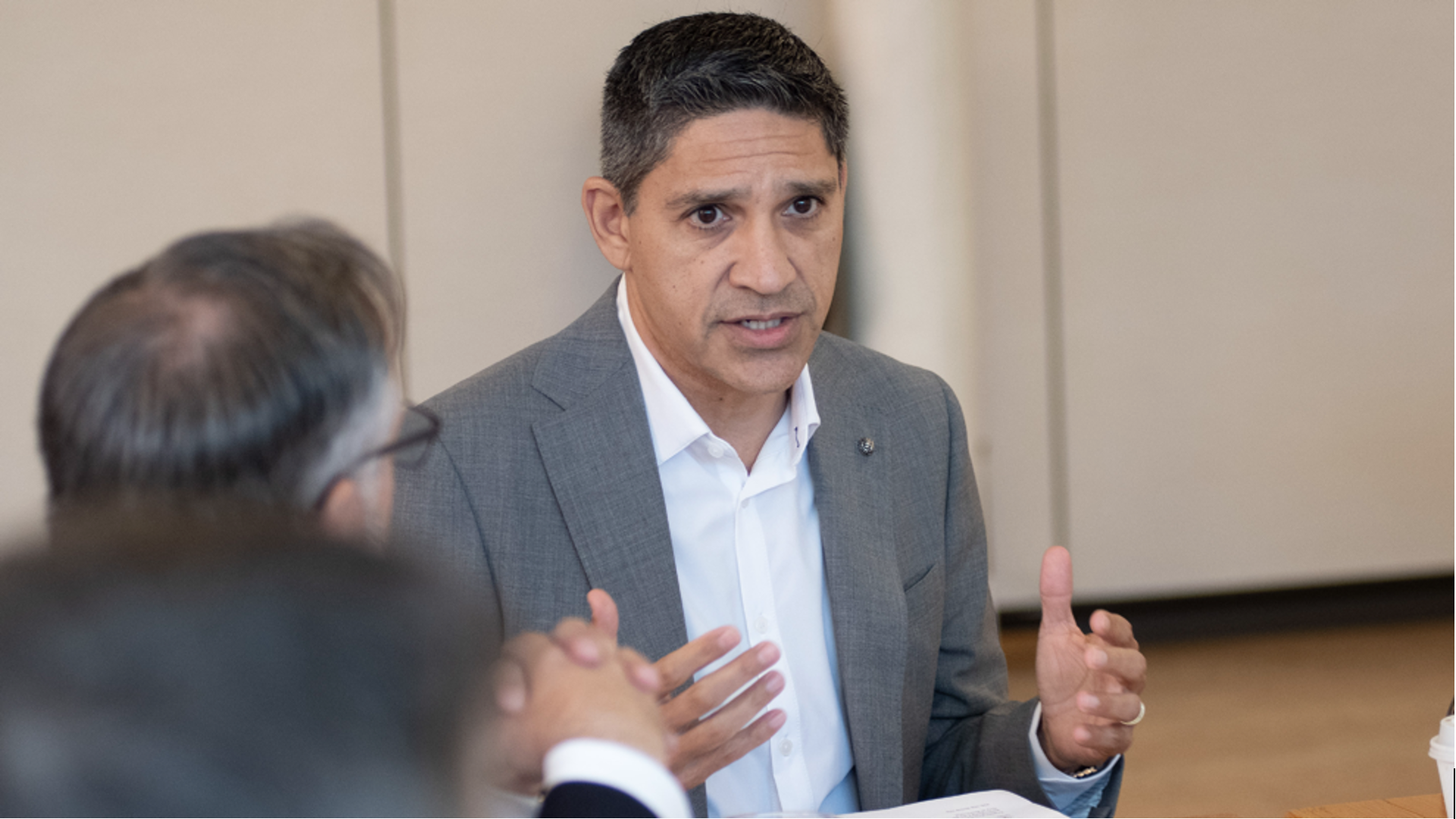 Man in a gray suit and white shirt speaking and gesturing with both hands during a meeting, with two blurred people in the foreground.