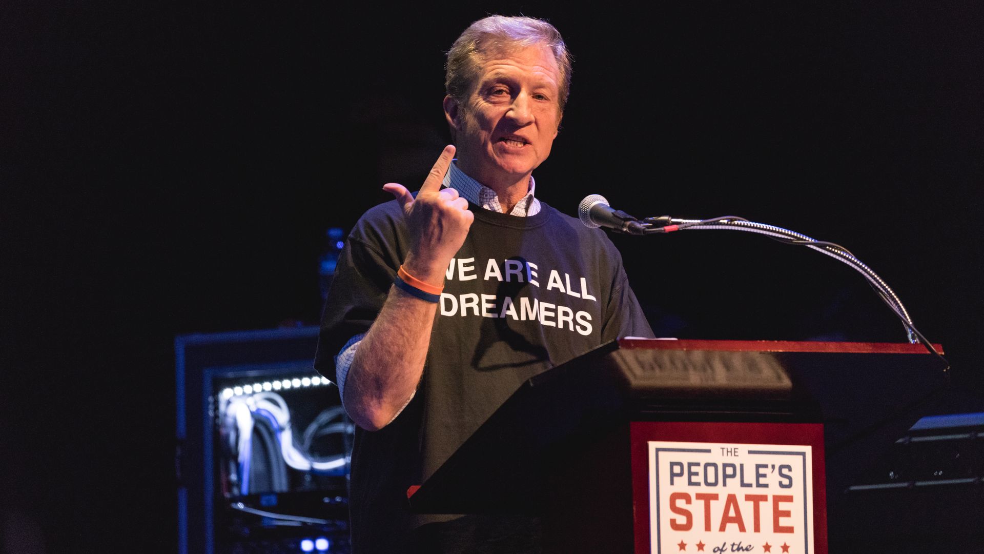 Tom Steyer standing at a podium with his finger in the air like he's counting