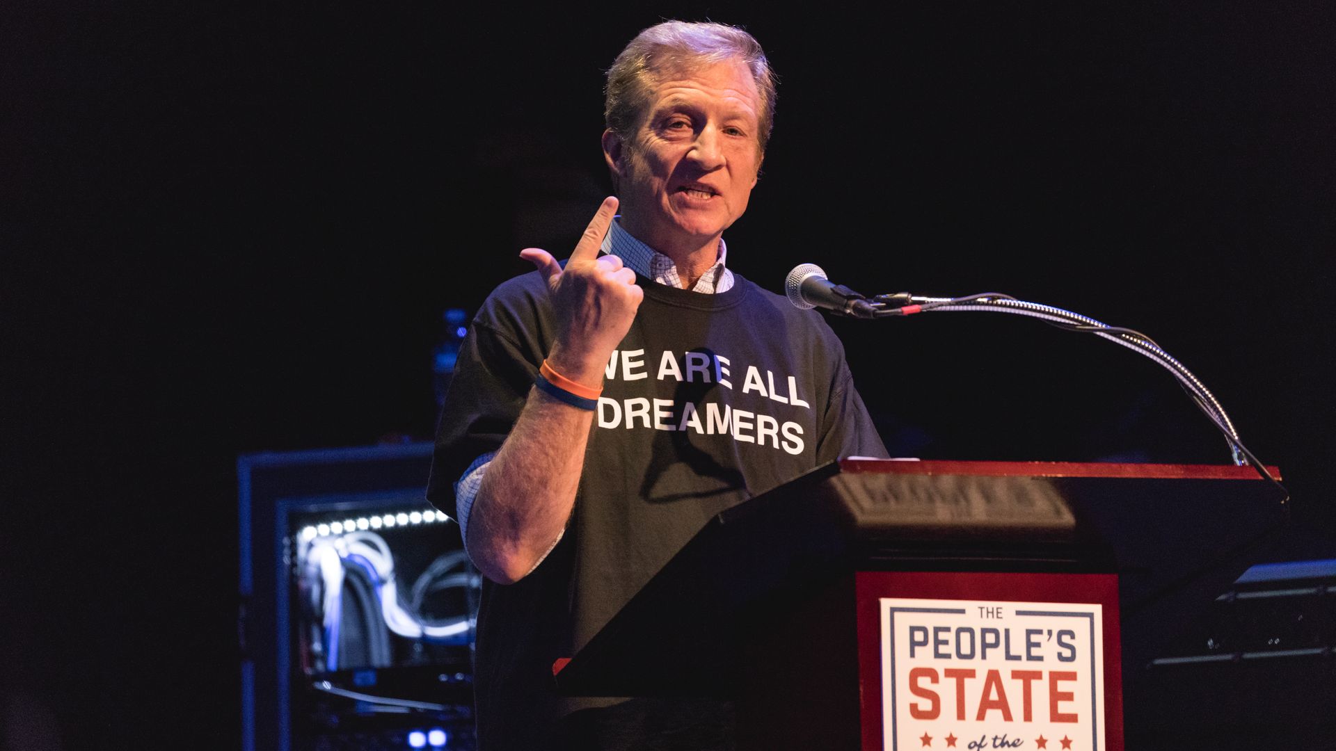 Tom Steyer standing at a podium with his finger in the air like he's counting