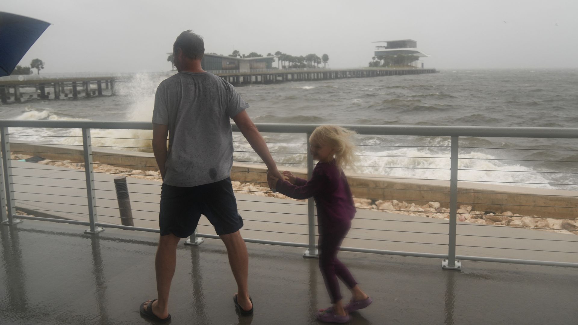 A man holds hands with a young girl as water splashes against a seawall. 