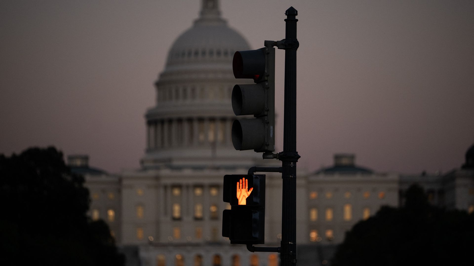 Orange hand pedestrian signal illuminated on a traffic light pole at dusk with the blurred U.S. Capitol building in the background under a dusky sky.