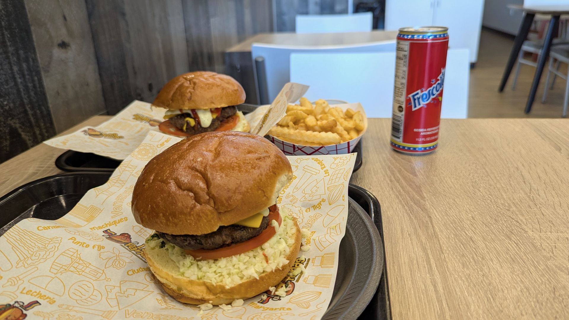 Two cheeseburgers on trays with a can of Frescolito on a table.
