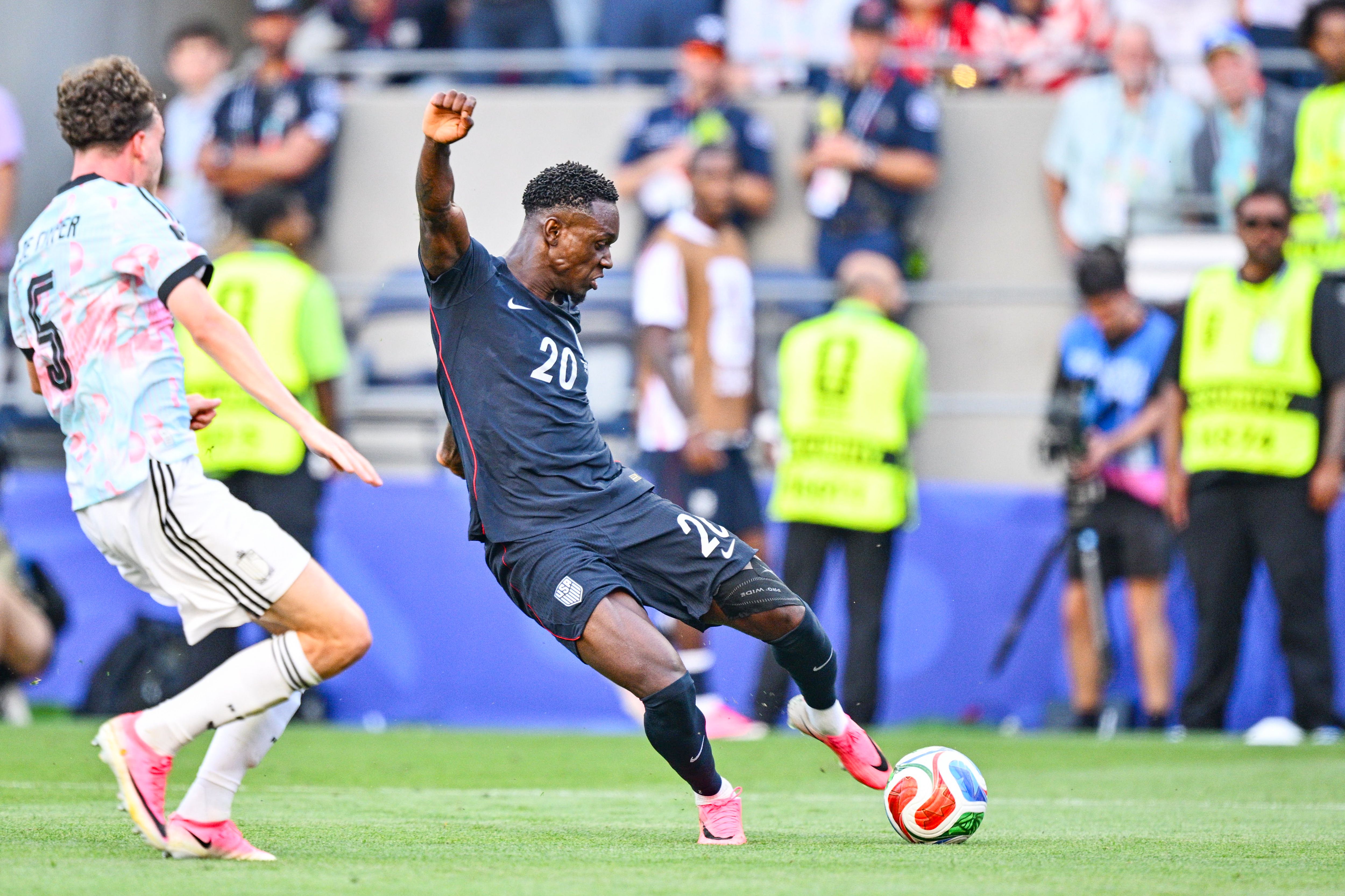 Folarin Balogun leands far to his right as his foot begins to kick a multicolored soccer ball, as a Belgian player approaches him and others can be seen blurry in the background.
