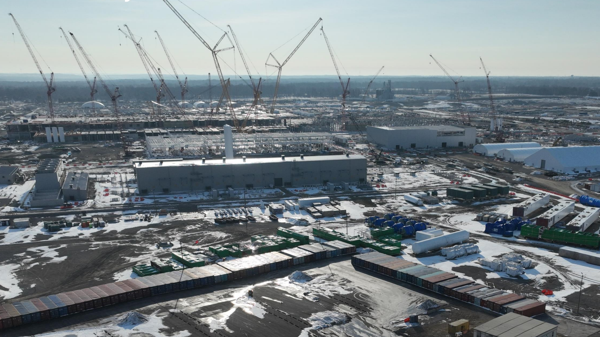 Aerial view of a snowy industrial construction site with multiple tall cranes, large white buildings, warehouses, stacked shipping containers, and snow-covered ground.