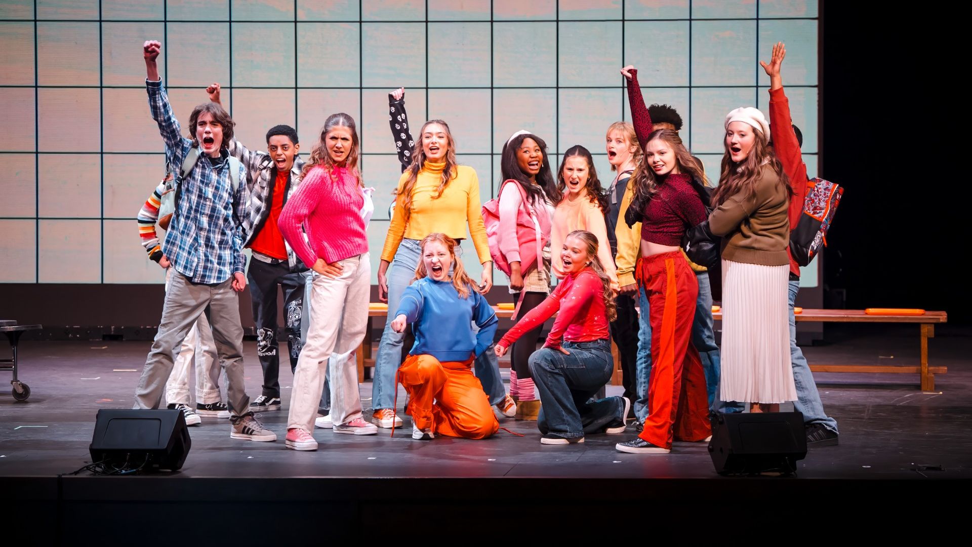 Group of diverse youths on a stage, cheering with raised arms. Colorful outfits (yellow, pink, blue, orange) contrast with a tiled wall backdrop.