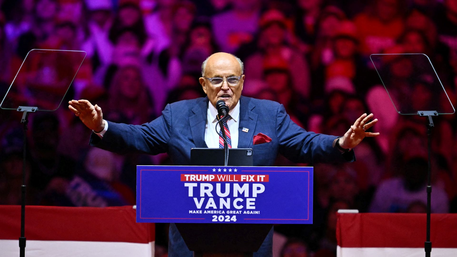 A bald-headed, bespectacled man, former New York City Mayor Rudy Giuliani, wearing a navy jacket, white shirt, and red, white and blue striped tie with white polka dots on stage in an arena at a lectern during a Trump rally, as people watch from behind under pink lights.