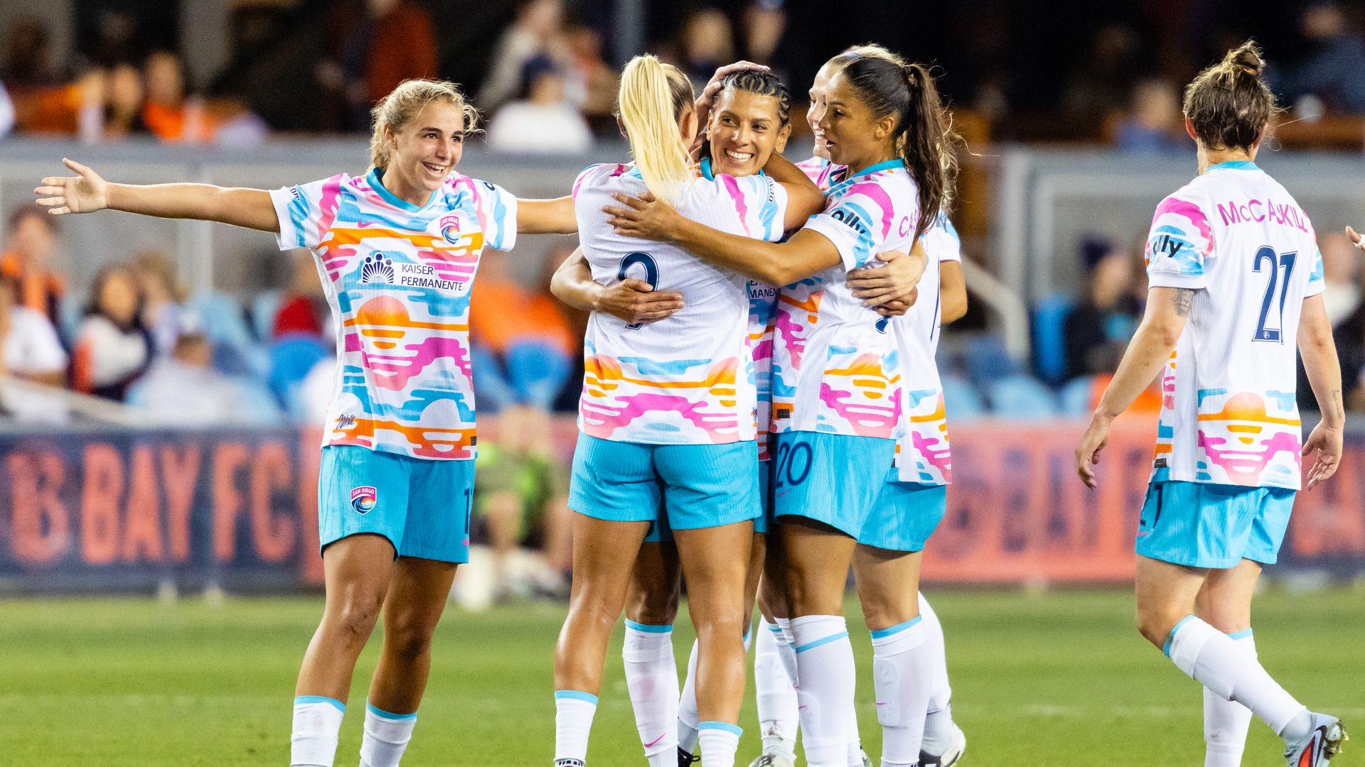 Female soccer players in colorful white jerseys with pink, orange, and blue patterns celebrate on the field with smiles and hugs during a game at night.