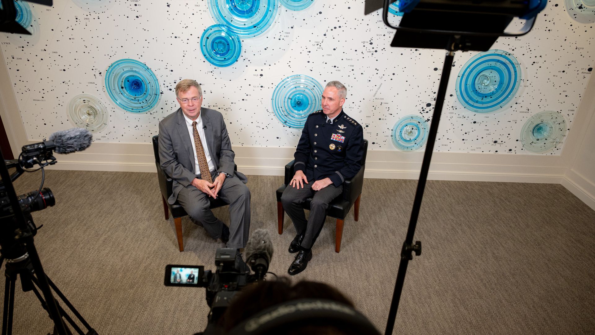 Two men seated for an interview; one in a gray suit and tie, the other in a decorated military uniform, with a white wall behind featuring blue concentric circle artwork.