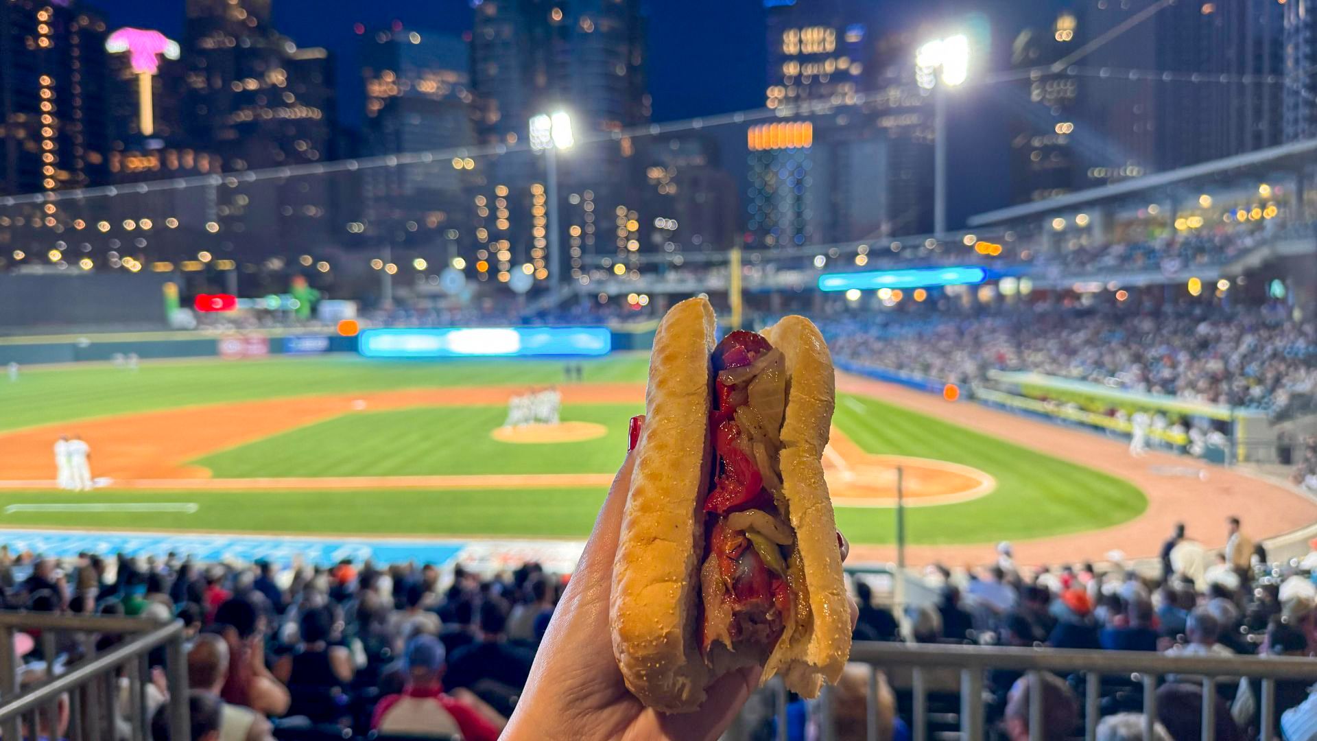 A hand holds a sausage sandwich topped with peppers and onions at a night baseball game, with a packed stadium and city skyline in the background.