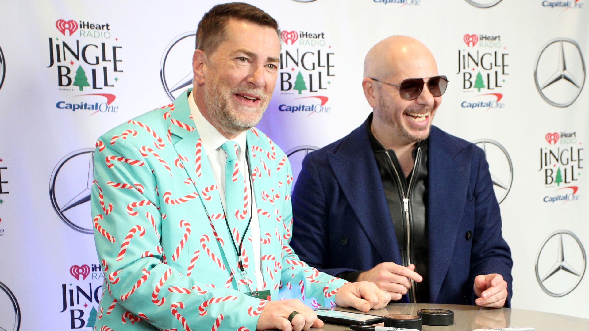 Two men sit at a round table in front of a Jingle Ball backdrop with iHeart logos; Dave Ryan wears a turquoise candy-cane suit, Pitbull wears a navy blazer with sunglasses; both smile.