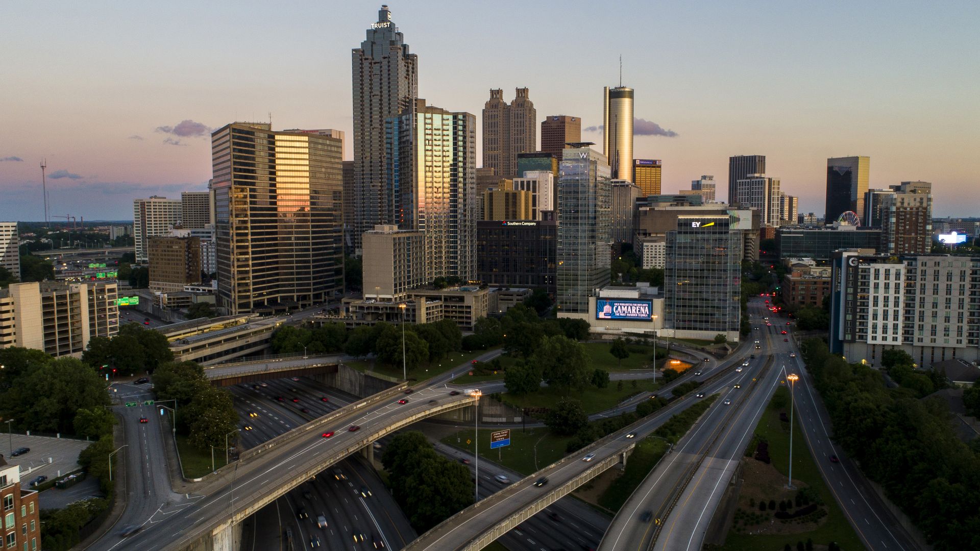 Aerial view of a city skyline at sunset with multiple highways and overpasses in the foreground, tall buildings reflecting orange and pink hues, and a clear sky.