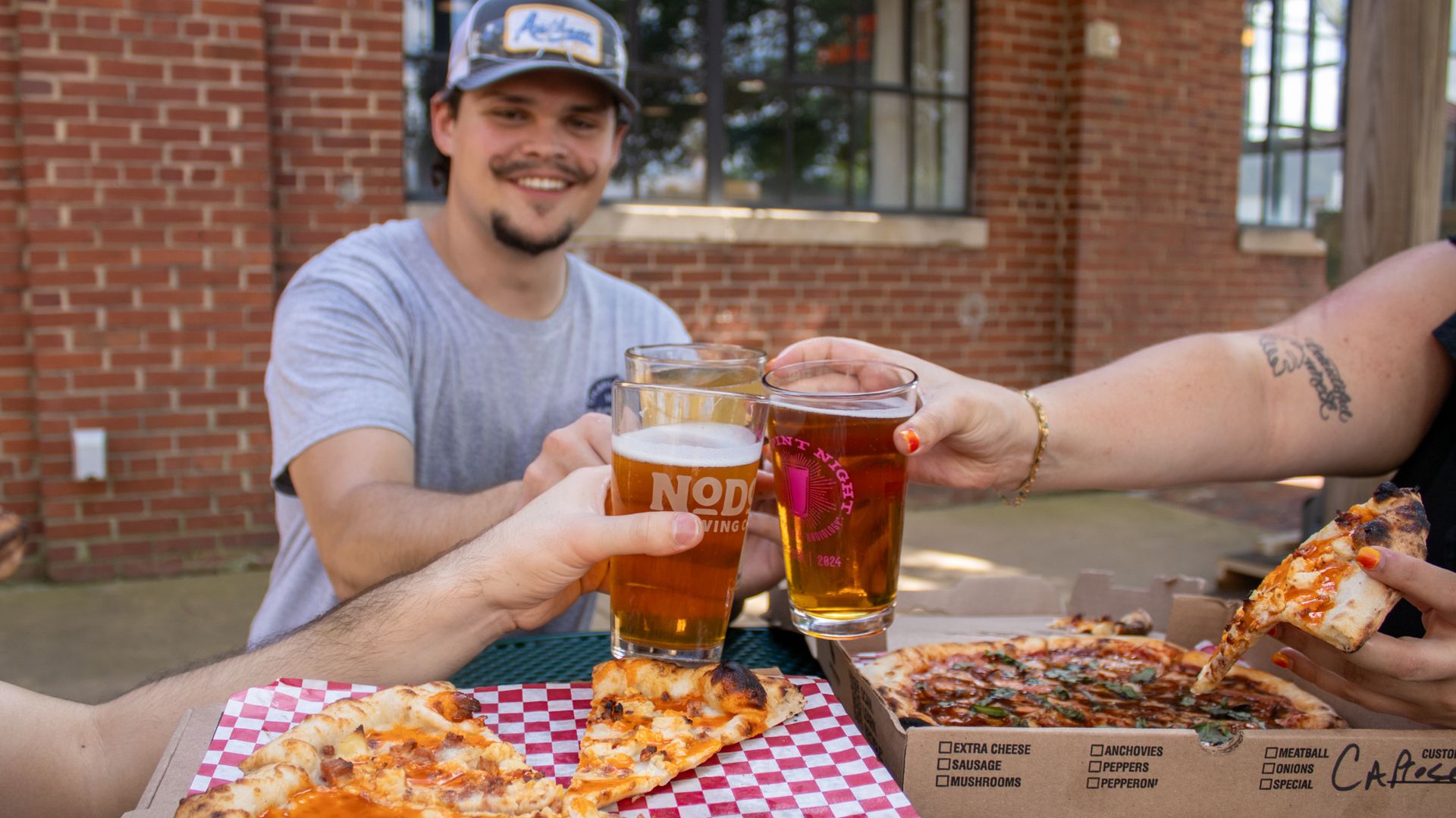 Group of friends outdoors toasting with glasses of beer over pizza on a green table, with brick building in background, one person holding a slice with a tattooed arm.