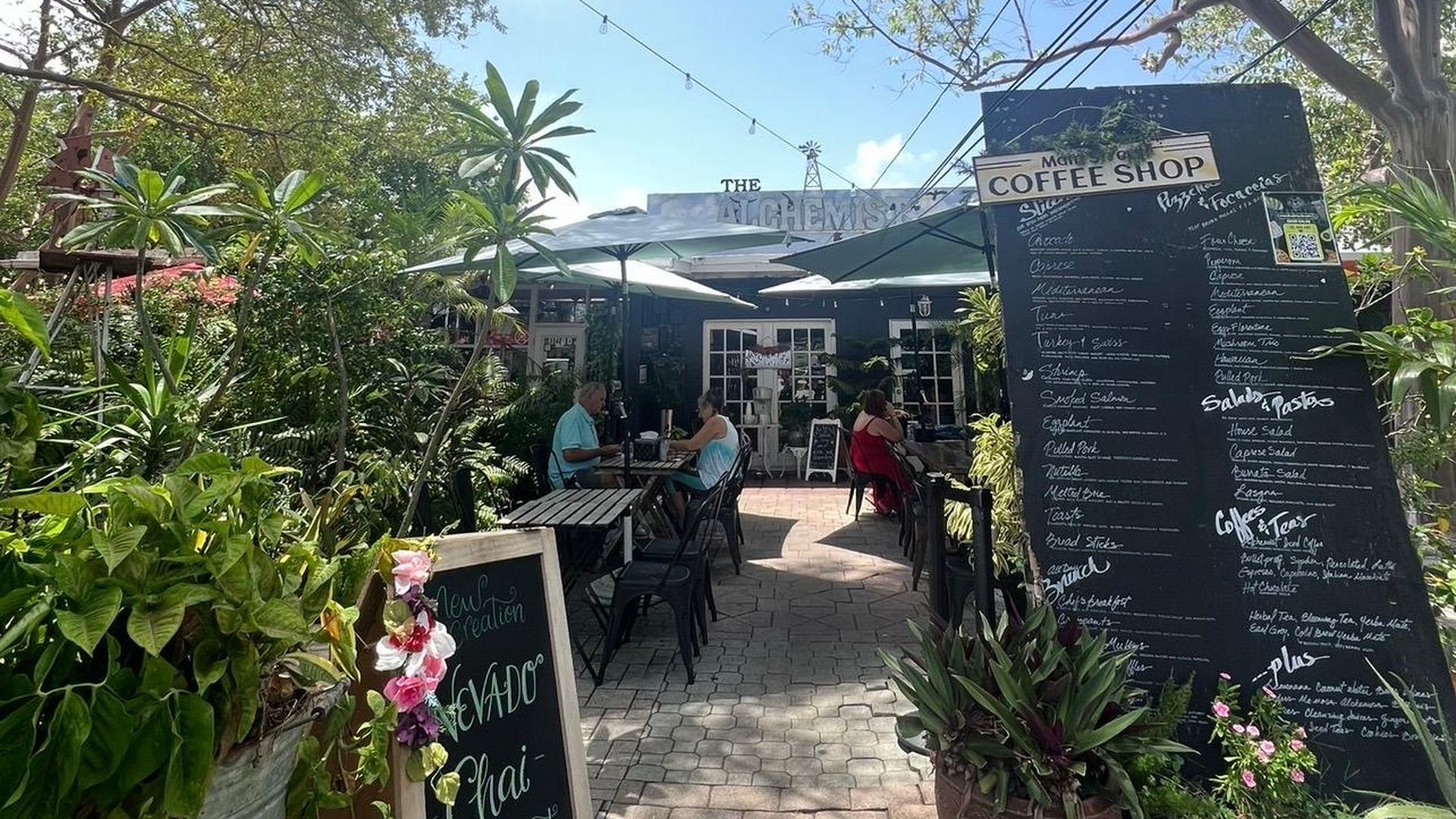 Outdoor cafe with chalkboard menus, lush potted plants, and string lights. Patrons sit at tables under umbrellas; tall trees frame a bright blue sky.