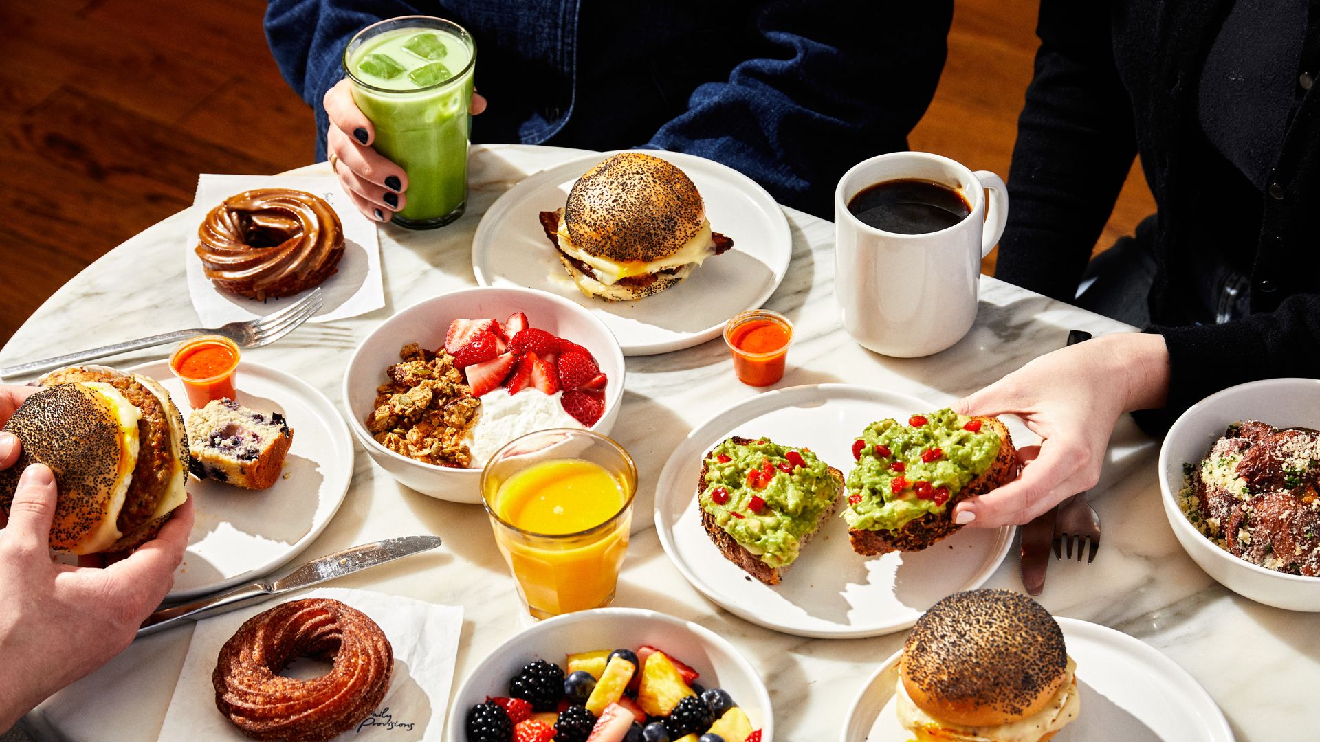 View of a breakfast table at Daily Provisions with poppy-seed bagels, an egg-and-cheese sandwich, avocado toast, bowls of fruit and granola, two doughnuts, a bright green smoothie, orange juice, and a white mug of coffee with one hand holding a breakfast sandwich and another grabbing avocado toast.