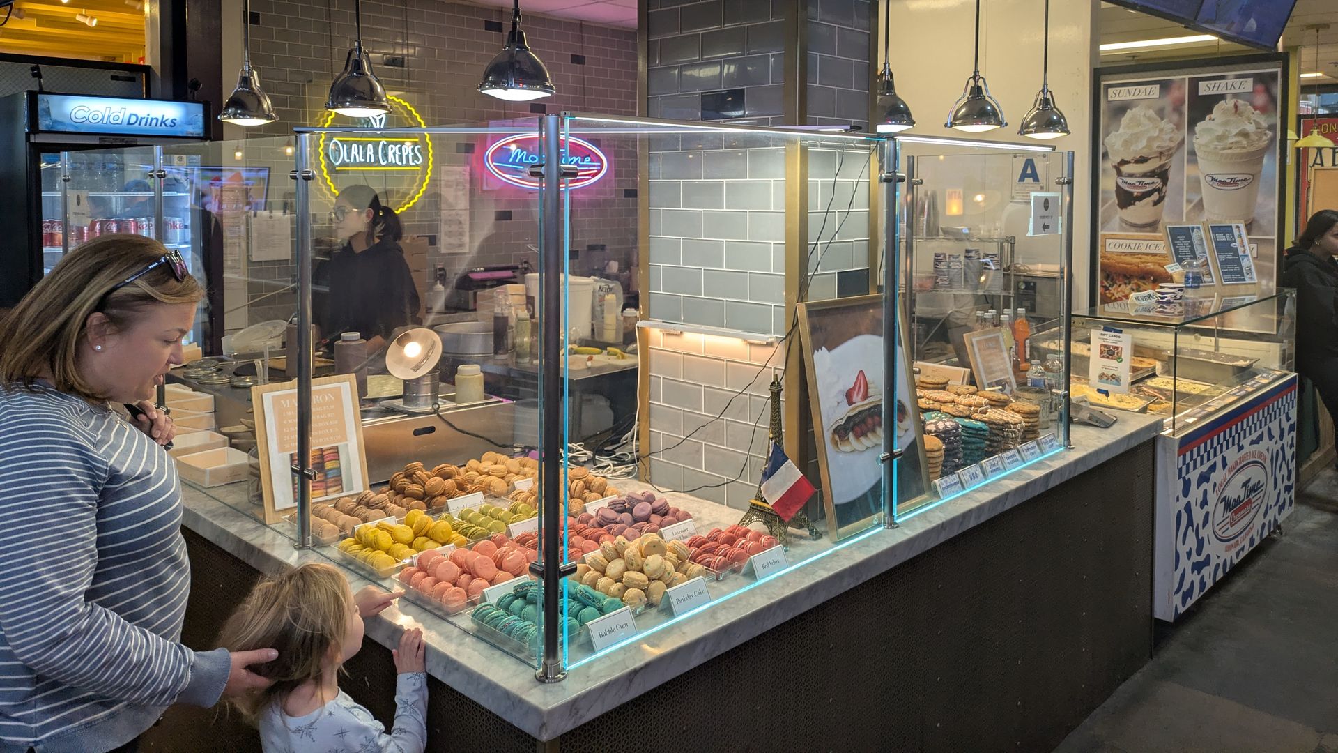 Woman and child looking at colorful macarons behind glass counter at a bakery stall with a menu above featuring croissants and crepes, inside a market.