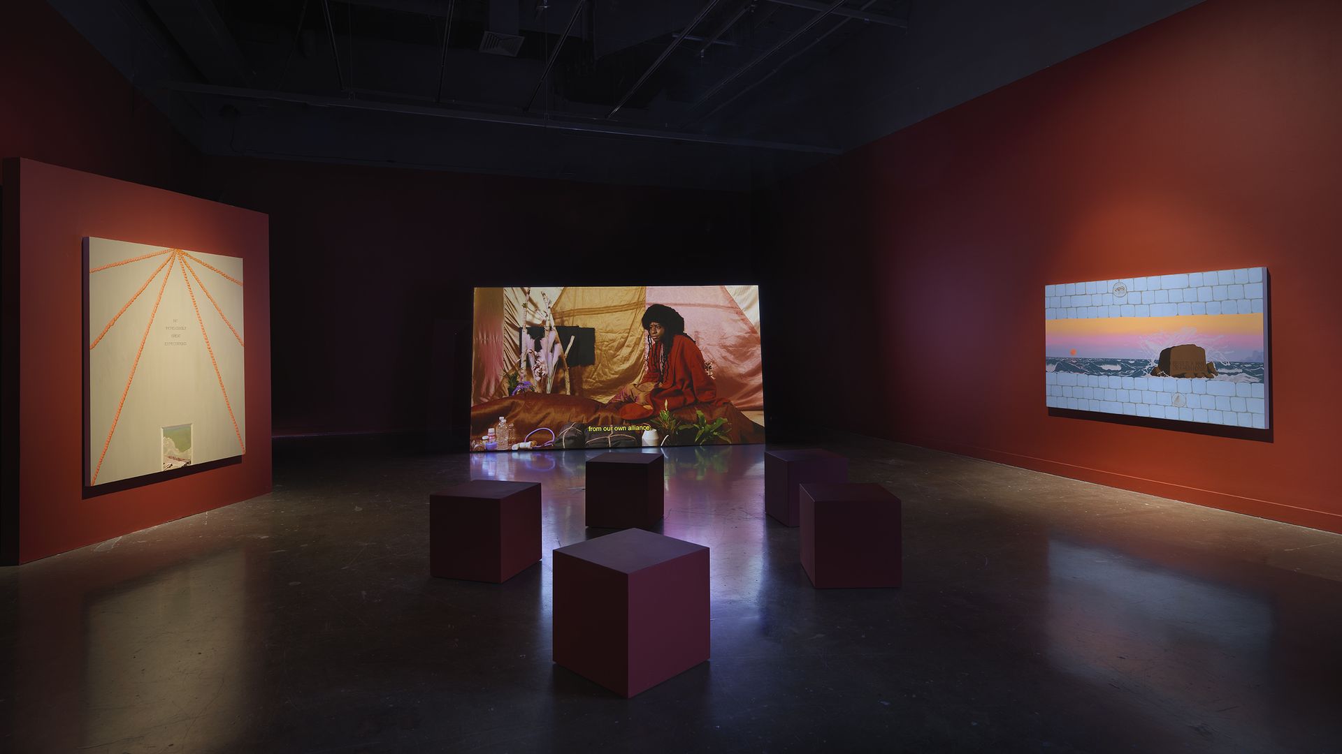Dim art gallery with red walls and an exposed dark ceiling. Colorful paintings hang along the walls, centered by a person in red; burgundy cube stools sit on the polished floor.