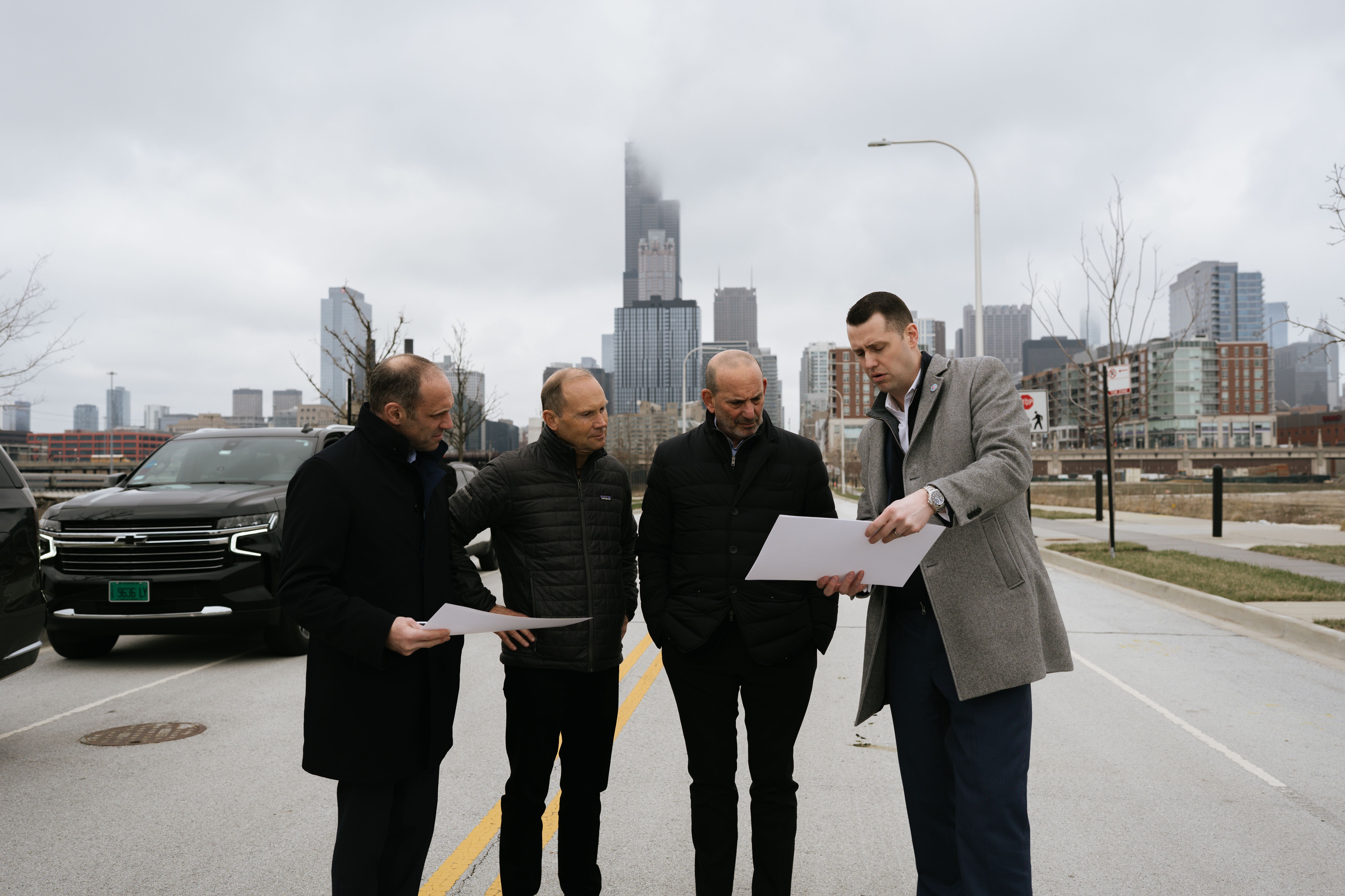 Photo of four men looking at a piece of paper on a street in front of a skyline. 