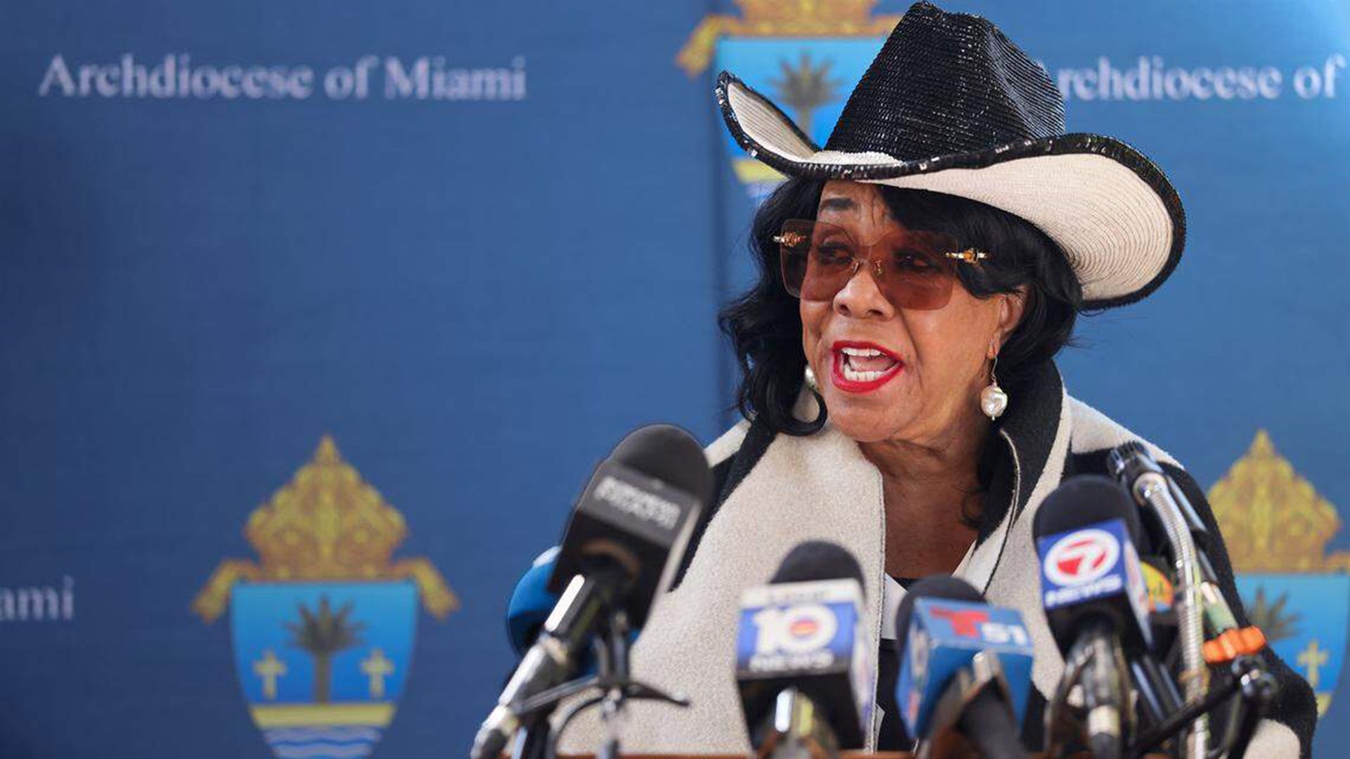 A woman wearing a black-and-white wide-brim hat and brown sunglasses speaks at a press conference, surrounded by multiple microphones, with a blue backdrop reading Archdiocese of Miami.