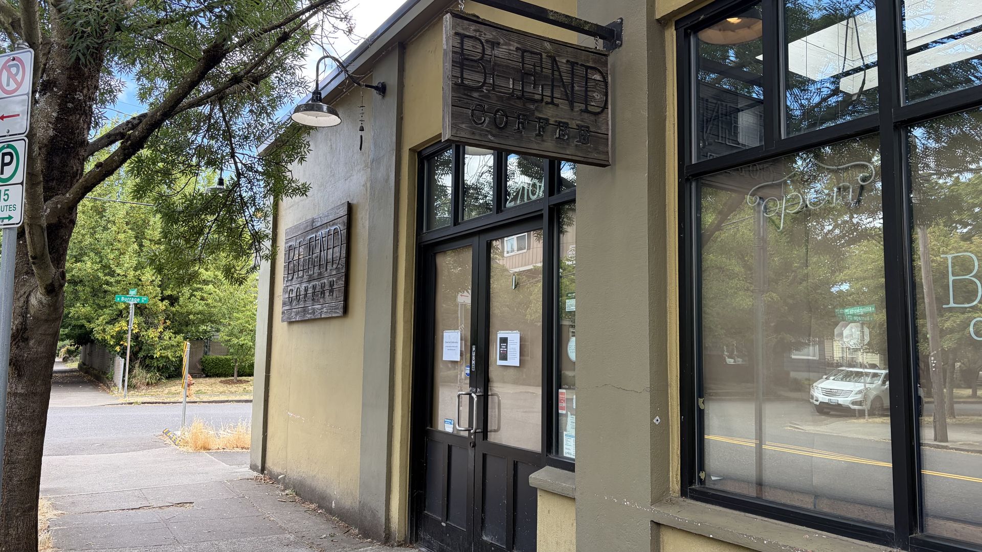 Sidewalk cafe exterior with a weathered wooden sign reading "BLEND COFFEE" above glass doors; a leafy tree, no-parking sign, and a car reflected in the storefront window.
