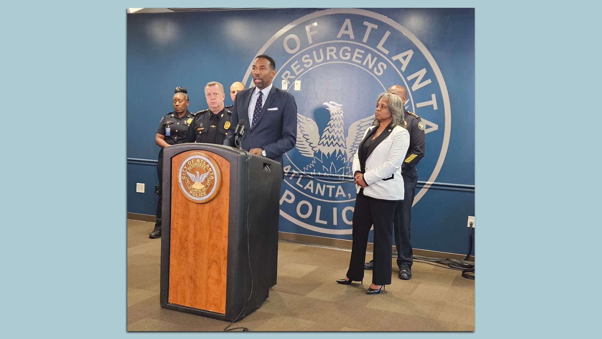 Mayor Andre Dickens speaking at city of Atlanta police podium with the city seal in the background. Several police officers and a woman in white blazer stand behind him in a press conference setting. 