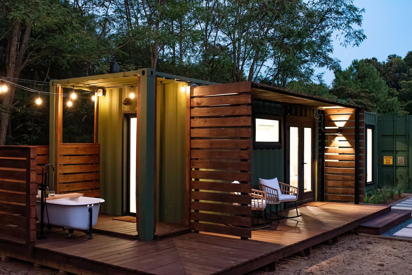 Outdoor evening view of a green shipping container home with wooden accents, warm string lights, a white clawfoot bathtub, and a cozy deck area with two white cushioned chairs.