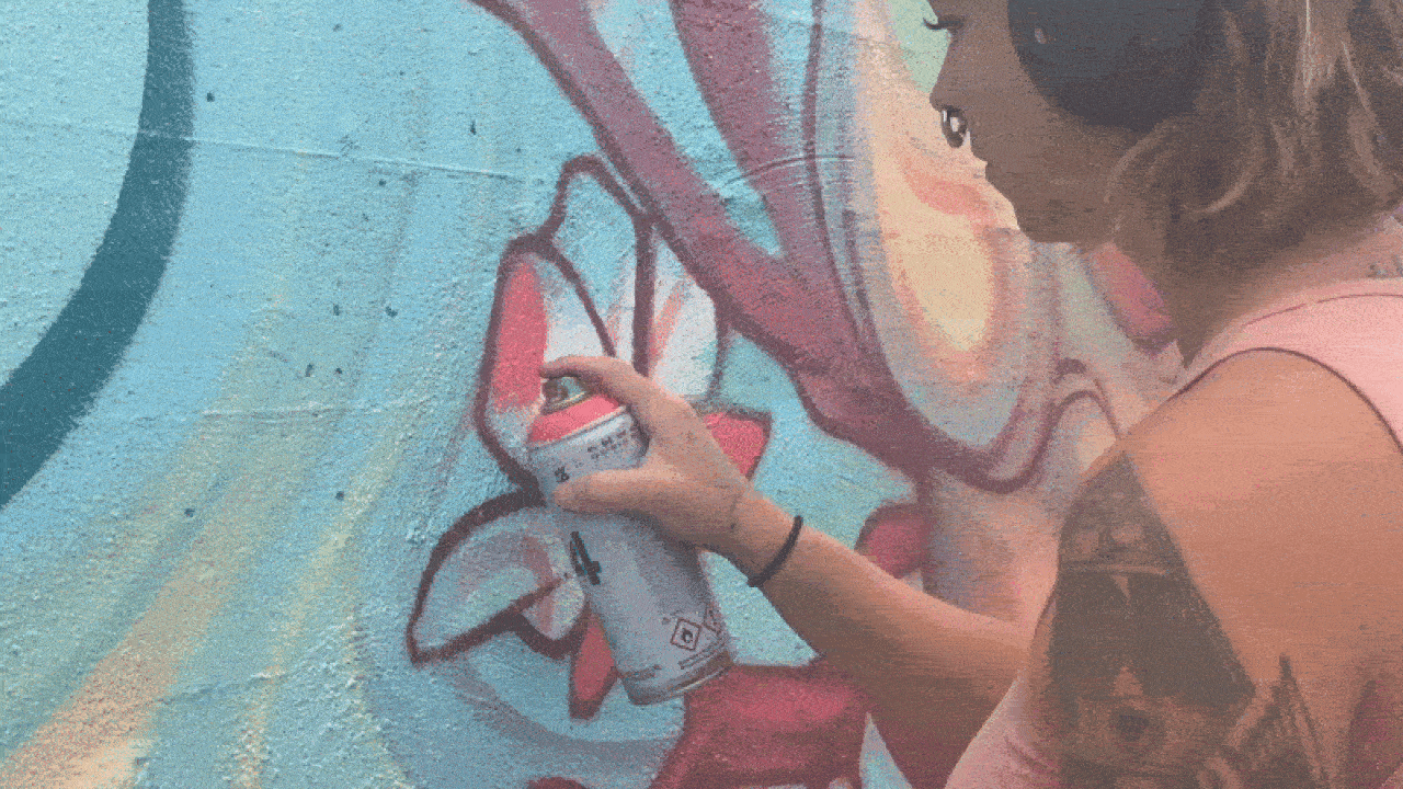 Person with tattooed arm spray painting red and white flower graffiti on a textured blue and yellow wall, wearing a pink sleeveless top.
