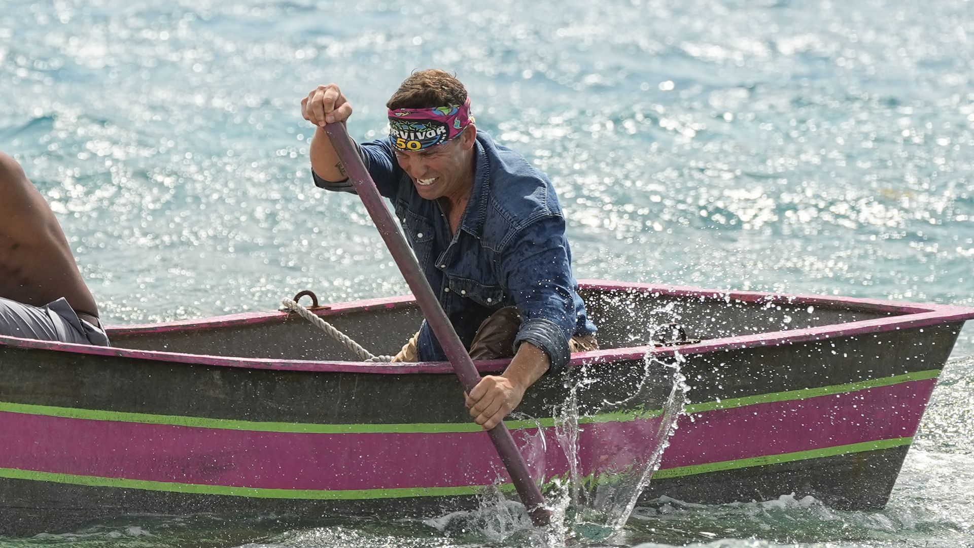Man wearing a colorful Survivor 50 headband and blue denim shirt paddling intensely in a pink and green boat on choppy water with splashes.