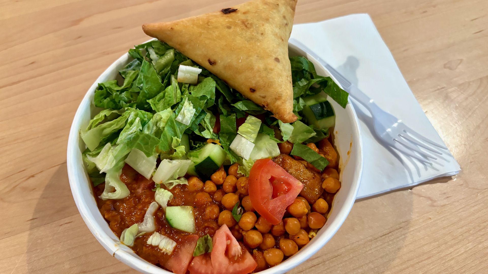 A white bowl topped with chickpeas, tomatoes, cucumbers, and shredded lettuce, plus a triangular sambusa; placed on a wooden table with a napkin and plastic fork.