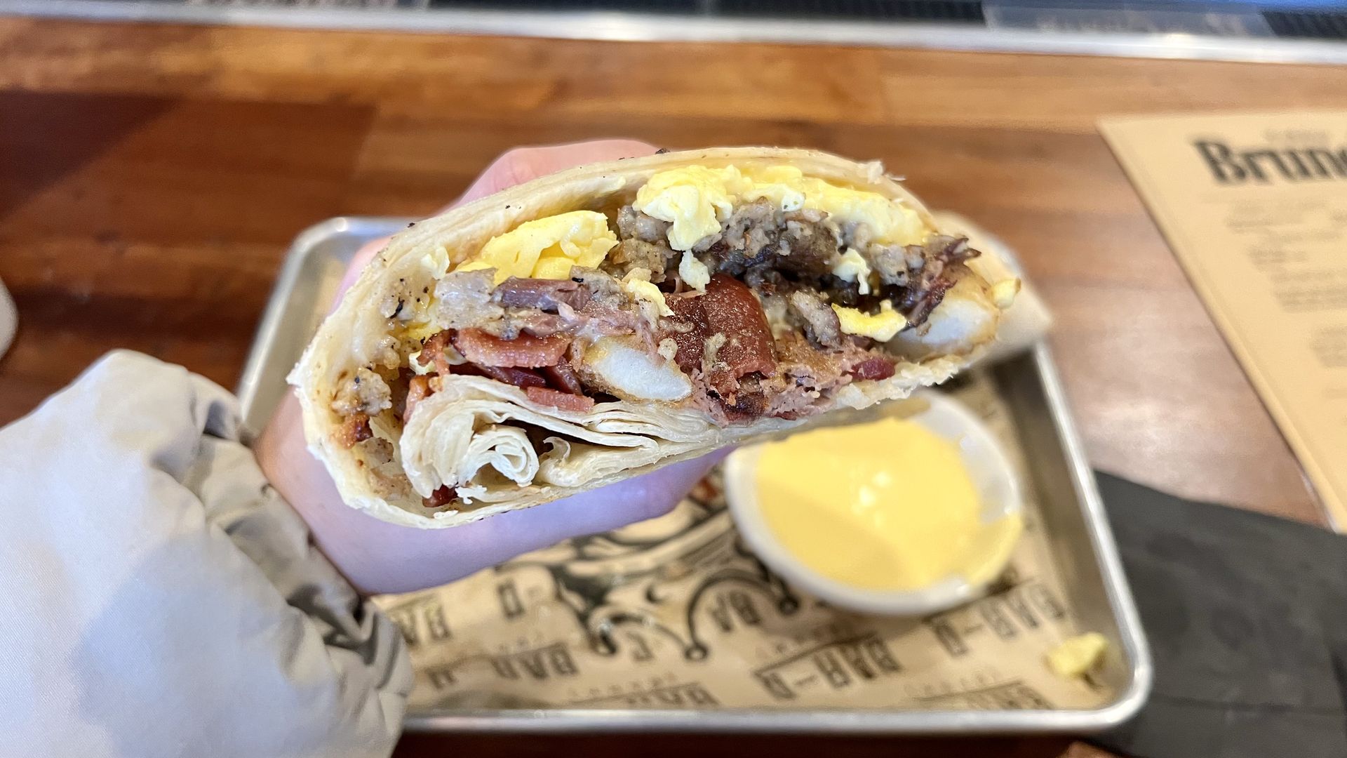 Hand holding a breakfast burrito with scrambled eggs, sausage, bacon, and potatoes, served on a metal tray with a side of yellow dipping sauce and a wooden table background.