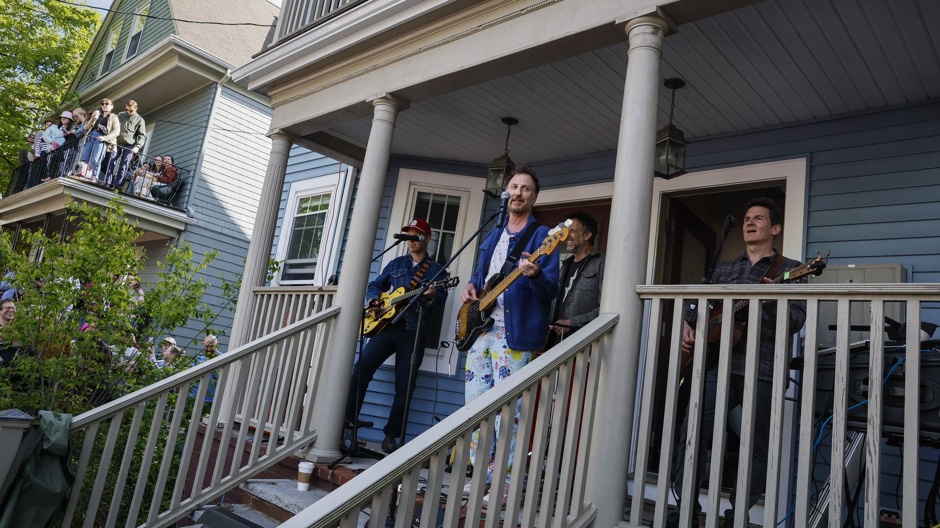 A live band performing on the porch of a blue house with white columns; three guitarists sing and play while a crowd watches from a balcony on the left.