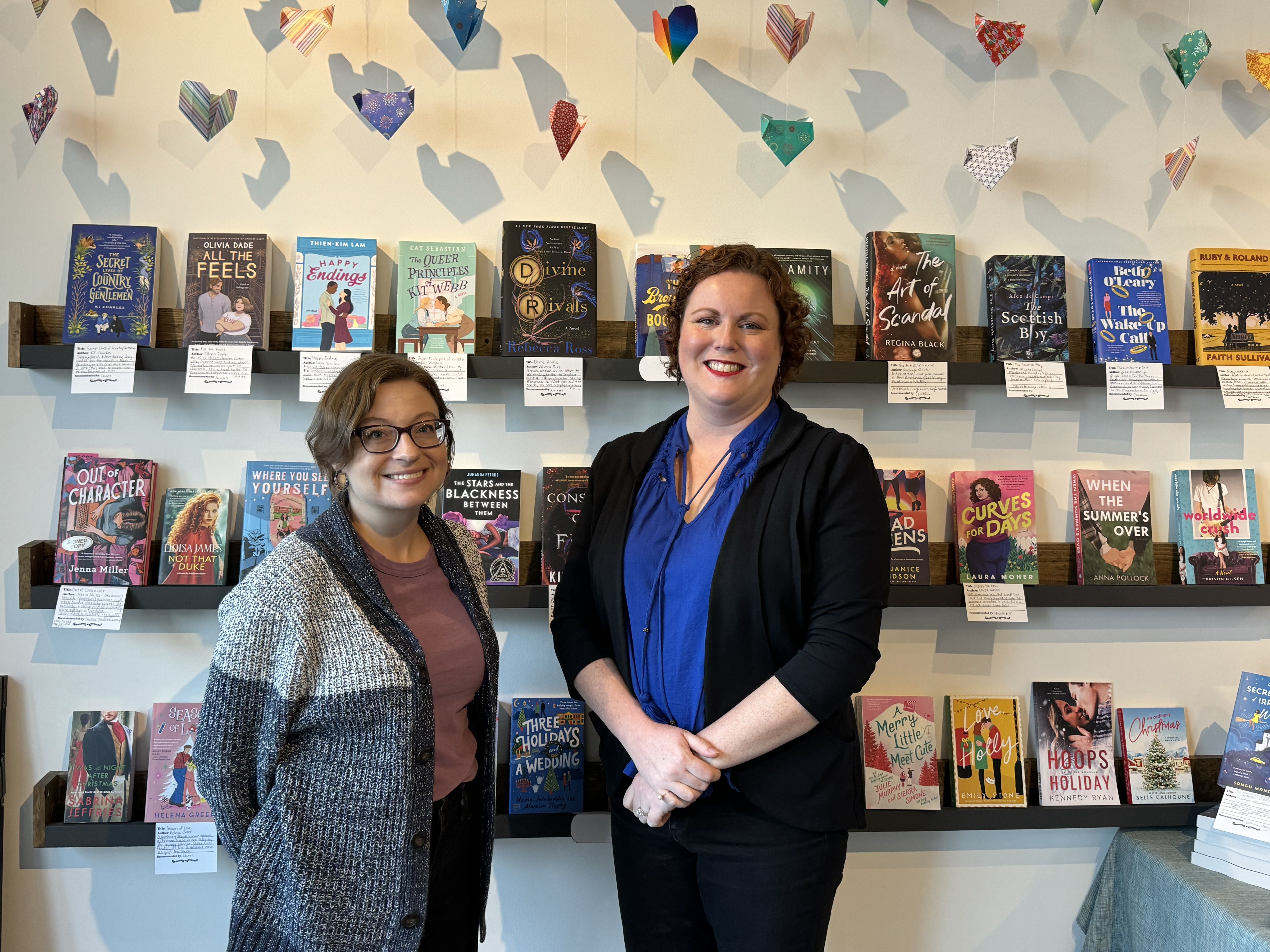 A woamn in a purple top and grey cardigan in glasses stands next to a taller woman with a blue shirt and black sweater in front of a row of books