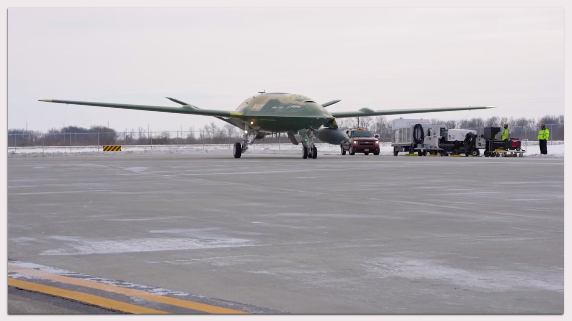 Green unmanned Navy aircraft on snowy airfield taxiway with ground crew in neon jackets, red truck, and equipment under gray sky.