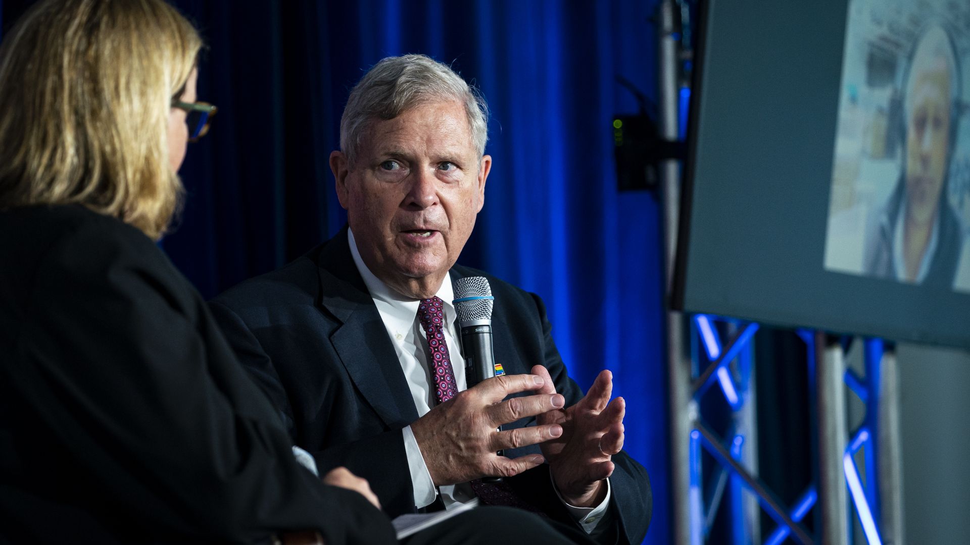 Agriculture Secretary Tom Vilsack, wearing a dark gray suit, white shirt and red tie, holds a microphone and speaks at an event.