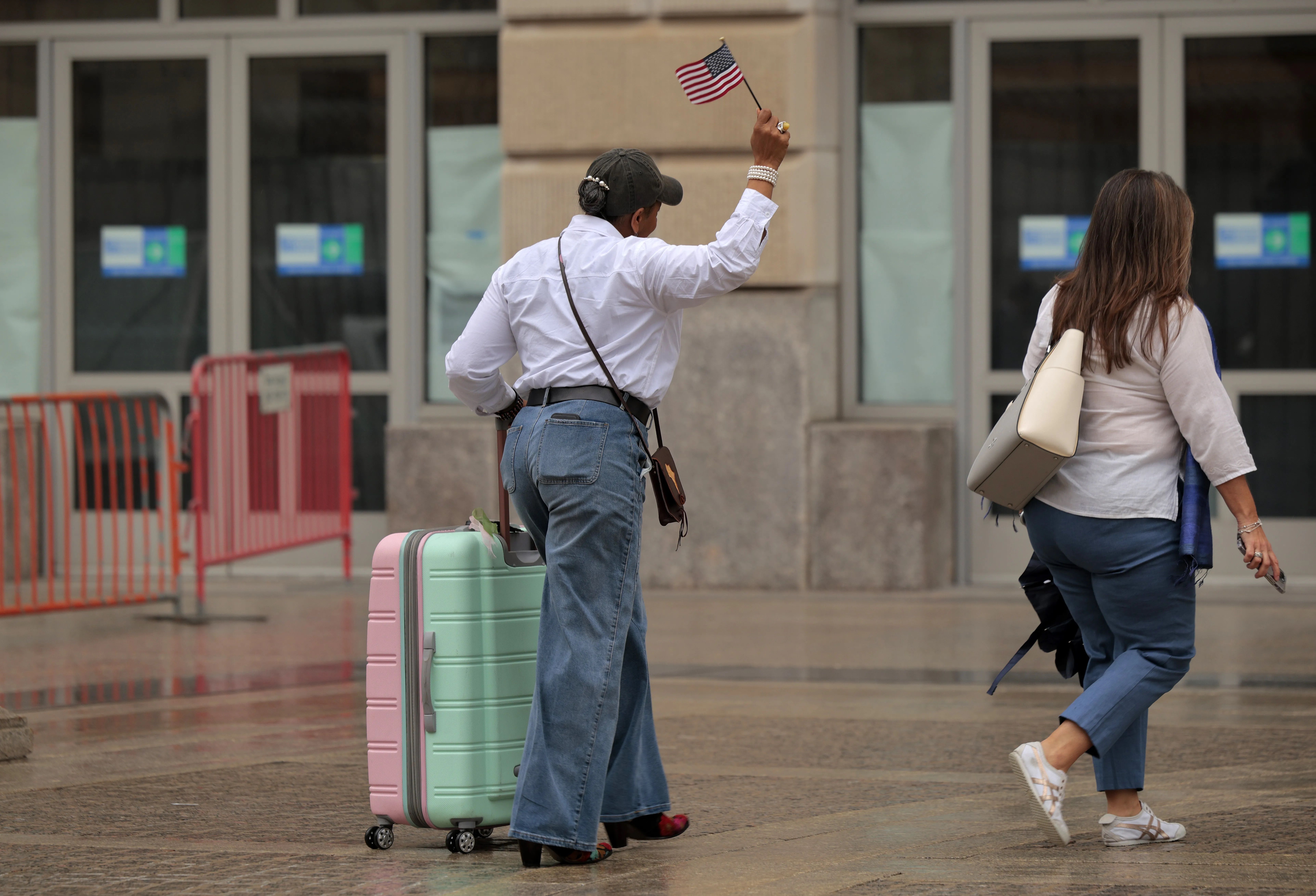 A person in a white shirt and blue jeans pulls a pastel pink and mint green suitcase, waving a small American flag, while another person walks ahead carrying a beige bag and wearing blue pants.