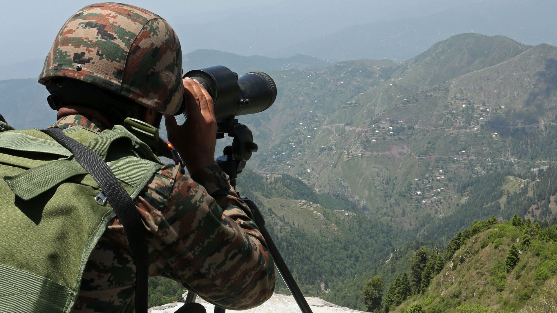 An Indian soldier looks through a monocular at the Line of Actual Control, a mountainous region in the distance. It is hazy.