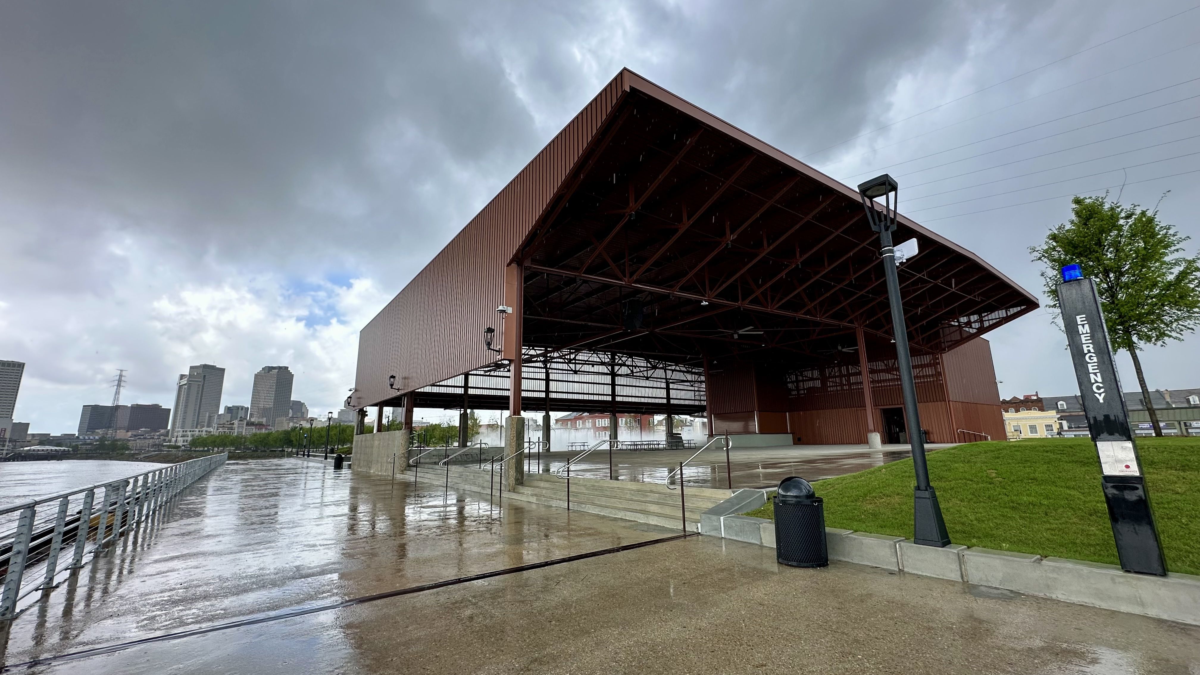 Stormy riverside with a large rust-colored open-air pavilion over a wet promenade. A few stairs and railing along the path; distant city skyline, green hill, and an emergency sign on the right.