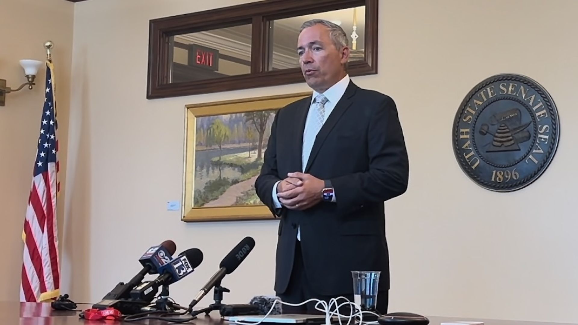 A man in a suit speaking in front of several microphones inside a government buliding with a flag.