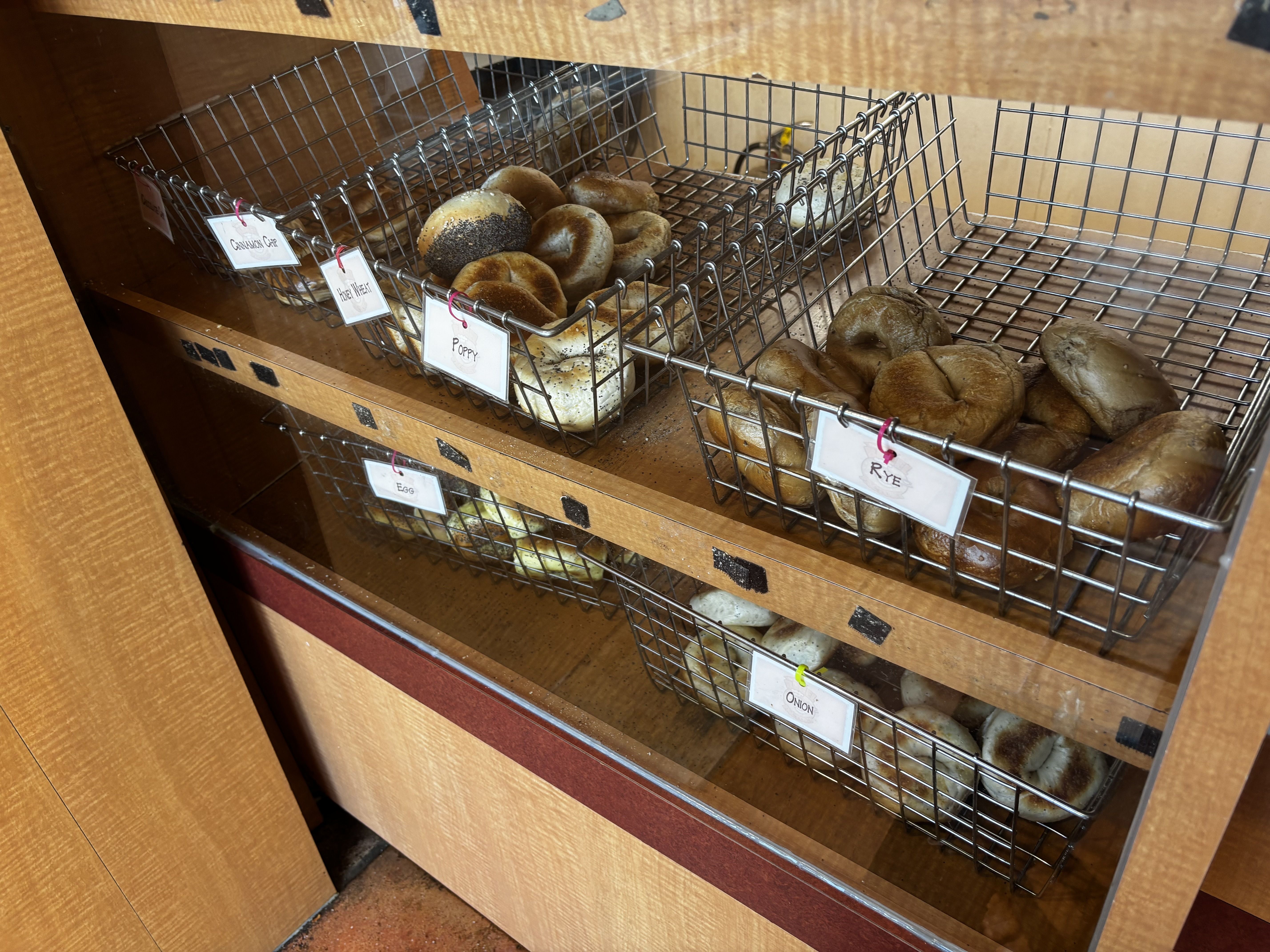 A bakery display of metal wire baskets filled with assorted bagels, including poppy, onion, and rye varieties, labeled with small white tags under a wooden shelf.
